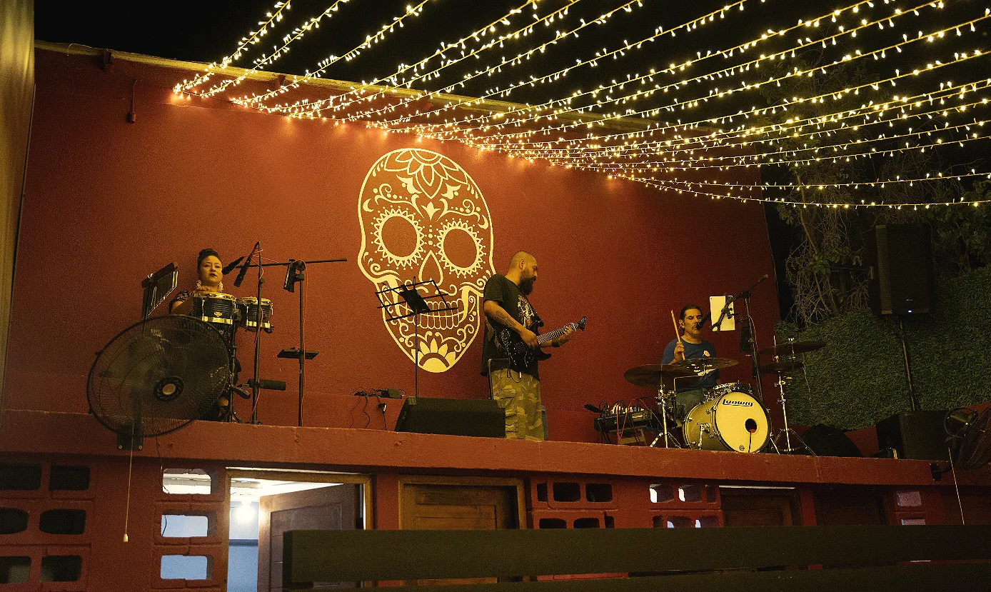 A trio playing on a red-walled stage under a canopy of string lights, with a painted calavera skull on the wall behind them