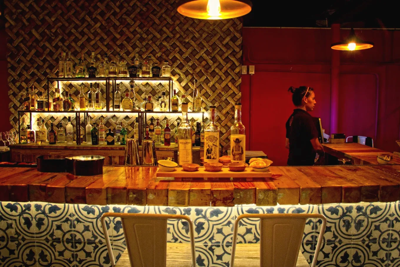 The bar at La Lupita: a wooden counter with blue-and-white tile, shelves of bottles lit from behind and a bartender in the background