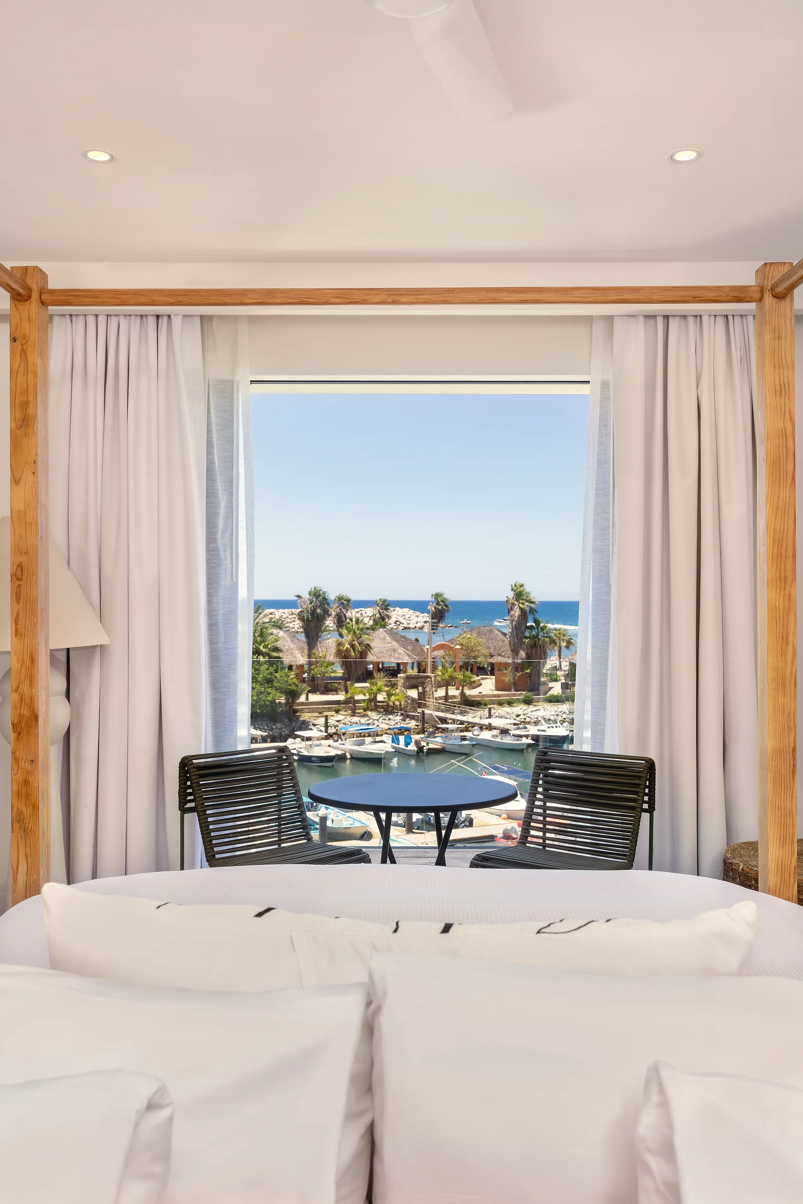 Bed with white linens in the foreground, framed by a timber four-post canopy, and a wide curtained glass opening beyond it onto a balcony with a small table and two chairs overlooking the marina and the sea.