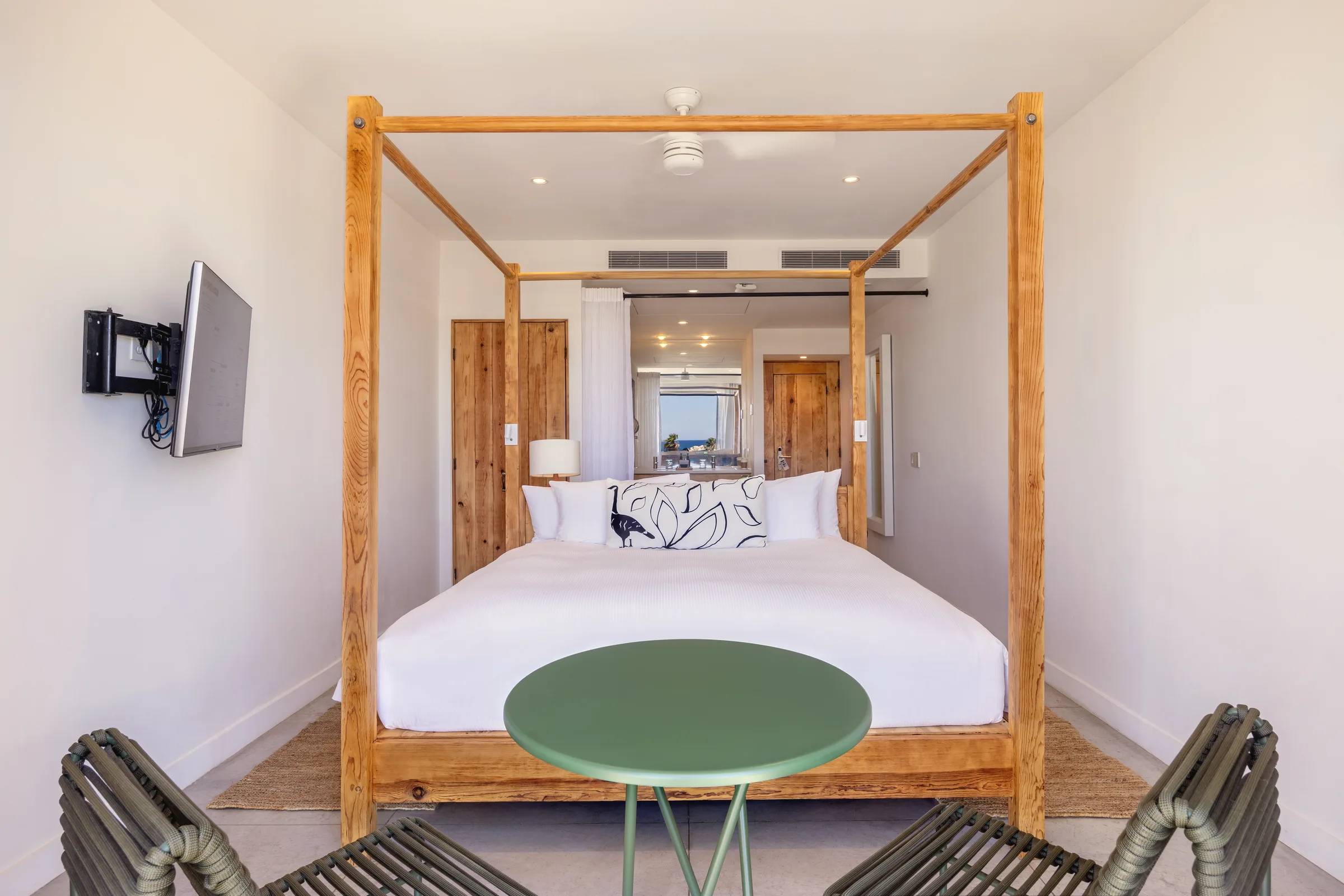Straight-on view of a king bed with a timber four-post canopy and white linens, a round green side table and two woven chairs in front, and wooden doors and a corridor behind.