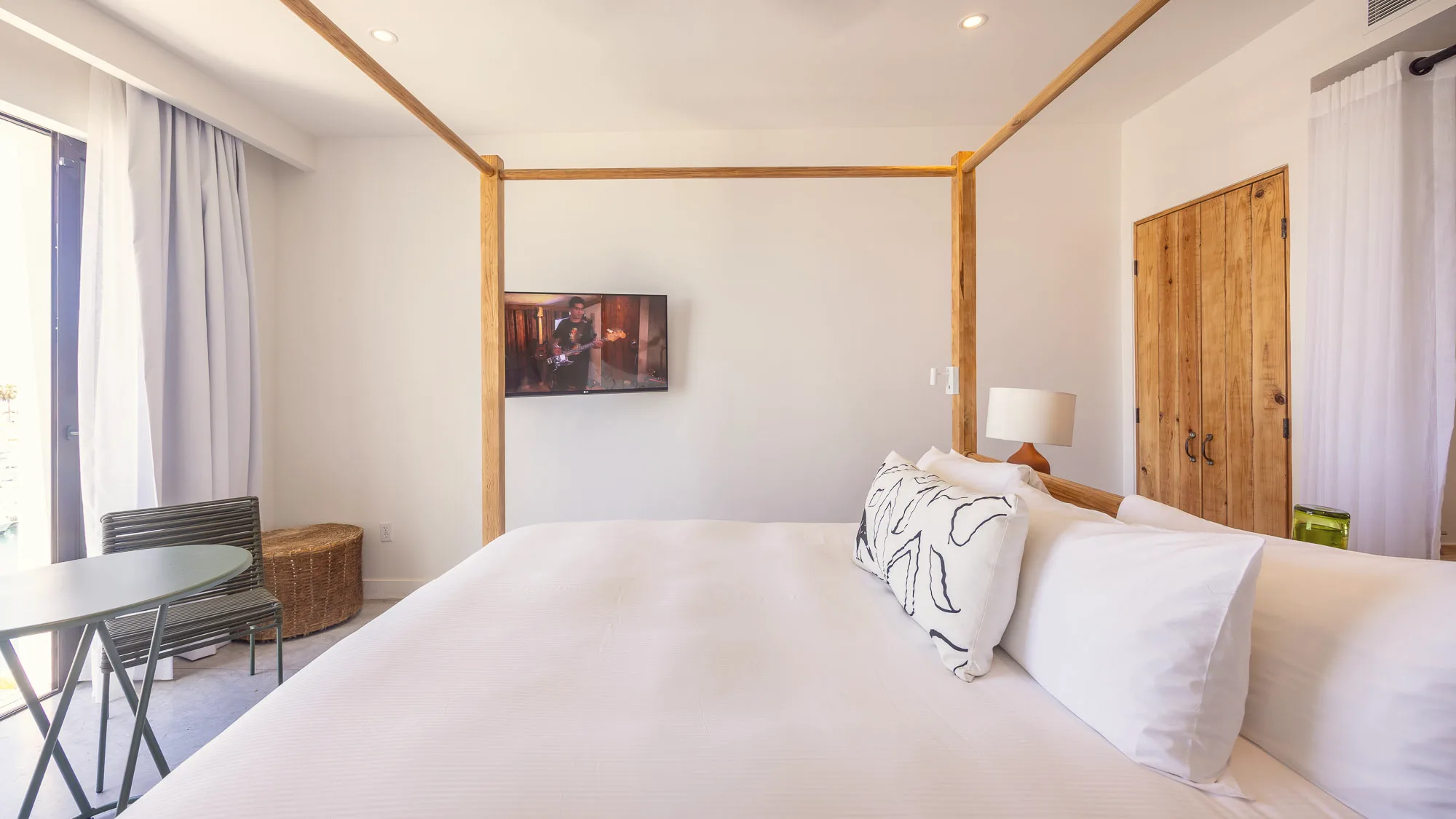 Side view of the king bed with white linens and patterned pillows under a timber canopy, with a wardrobe, a wall-mounted television and a round table and chair by the window.