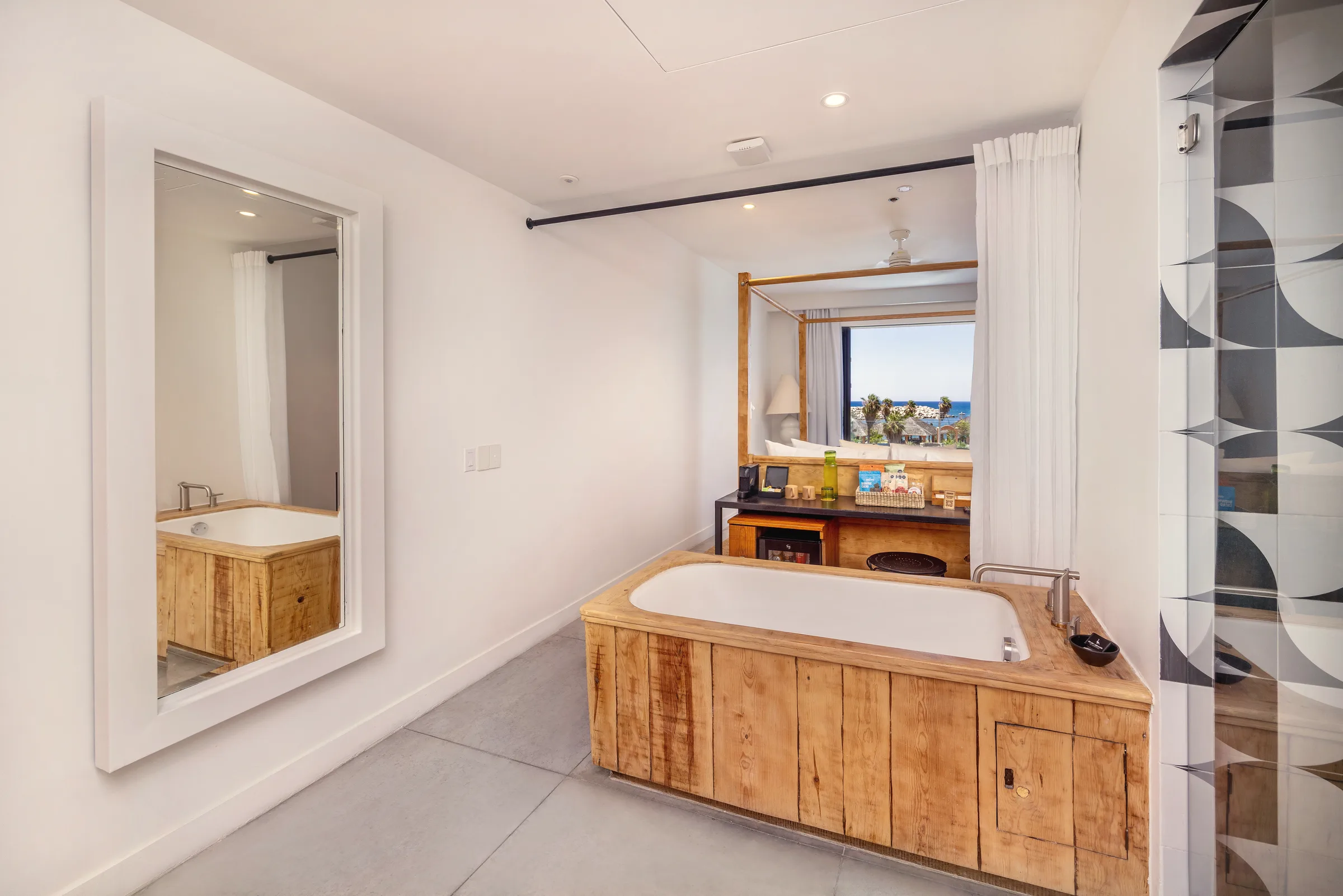 Bathroom area open to the bedroom, with a wooden soaking tub, a large white-framed mirror, a curtain, and a window showing palms and the sea beyond the bed.