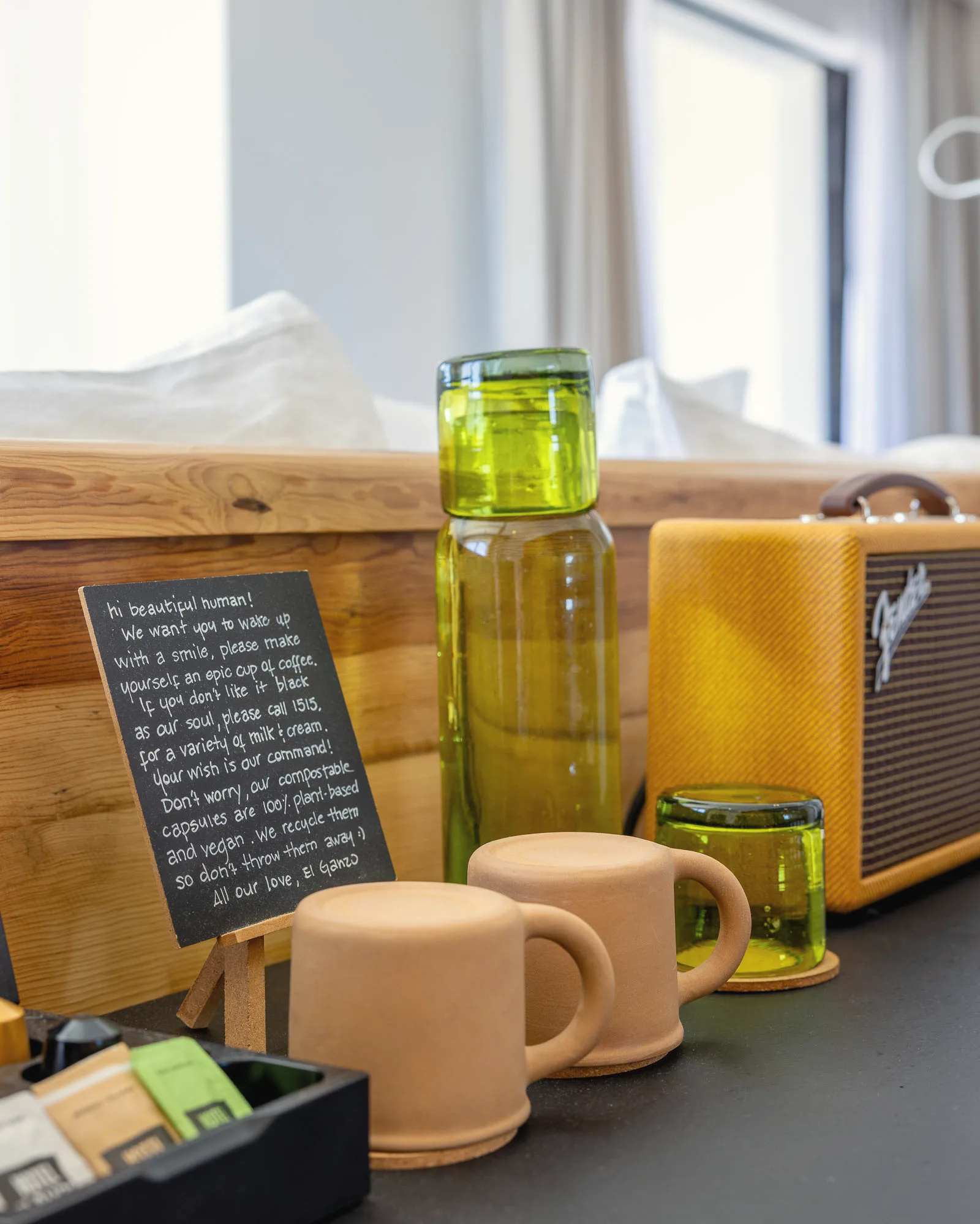 Close-up of a coffee tray with terracotta cups, a green glass carafe and tumbler, a yellow portable speaker and a handwritten chalkboard note, with the bed softly out of focus behind.