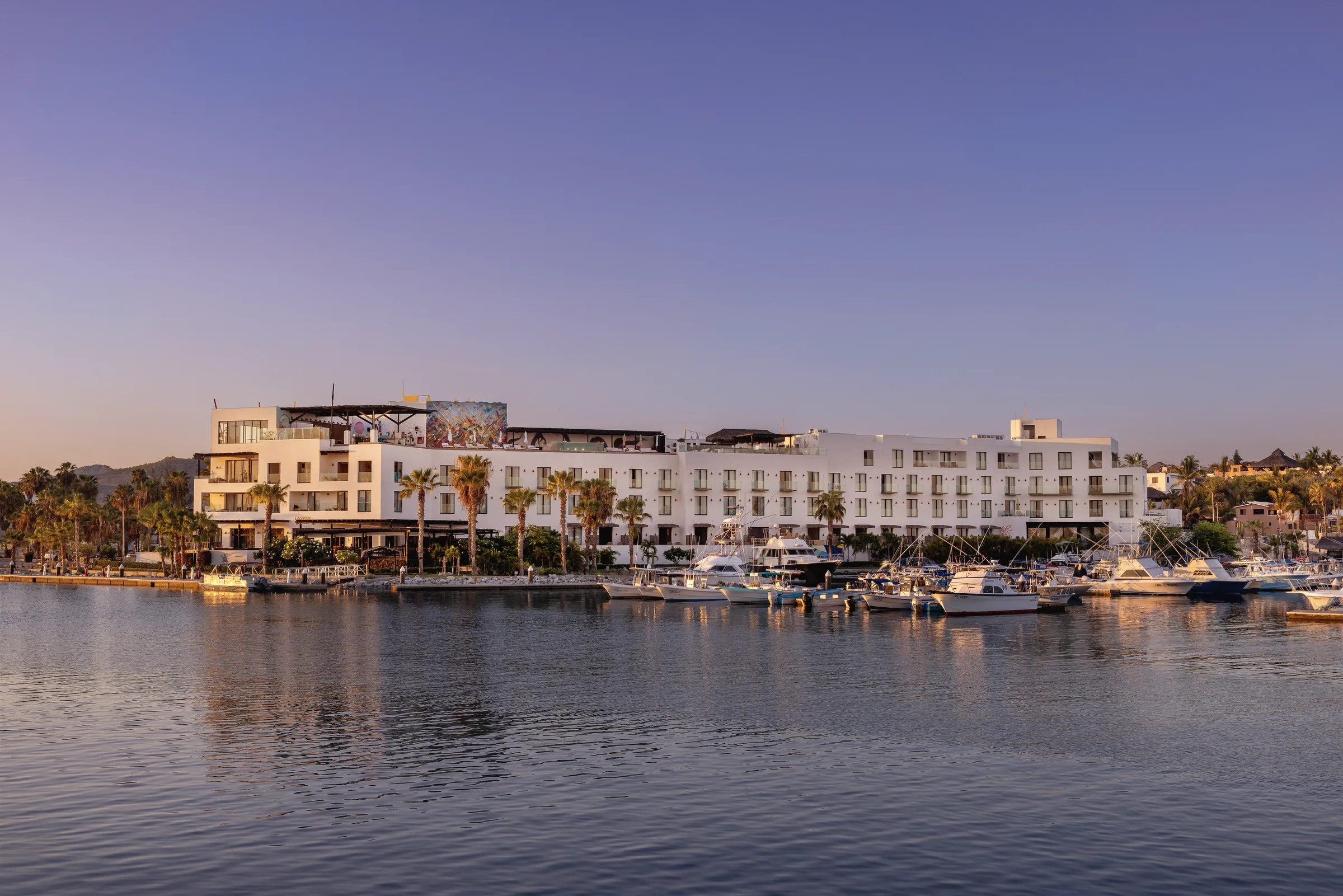 Hotel El Ganzo seen from across the water in warm low light, with boats moored along the marina in the foreground.