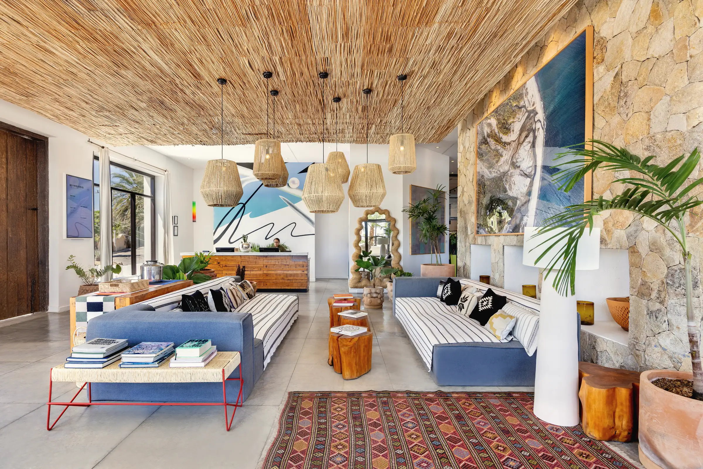 Hotel lobby with a woven-reed ceiling, woven pendant lamps, blue sofas, a patterned rug and an abstract blue-and-black mural behind the reception desk.