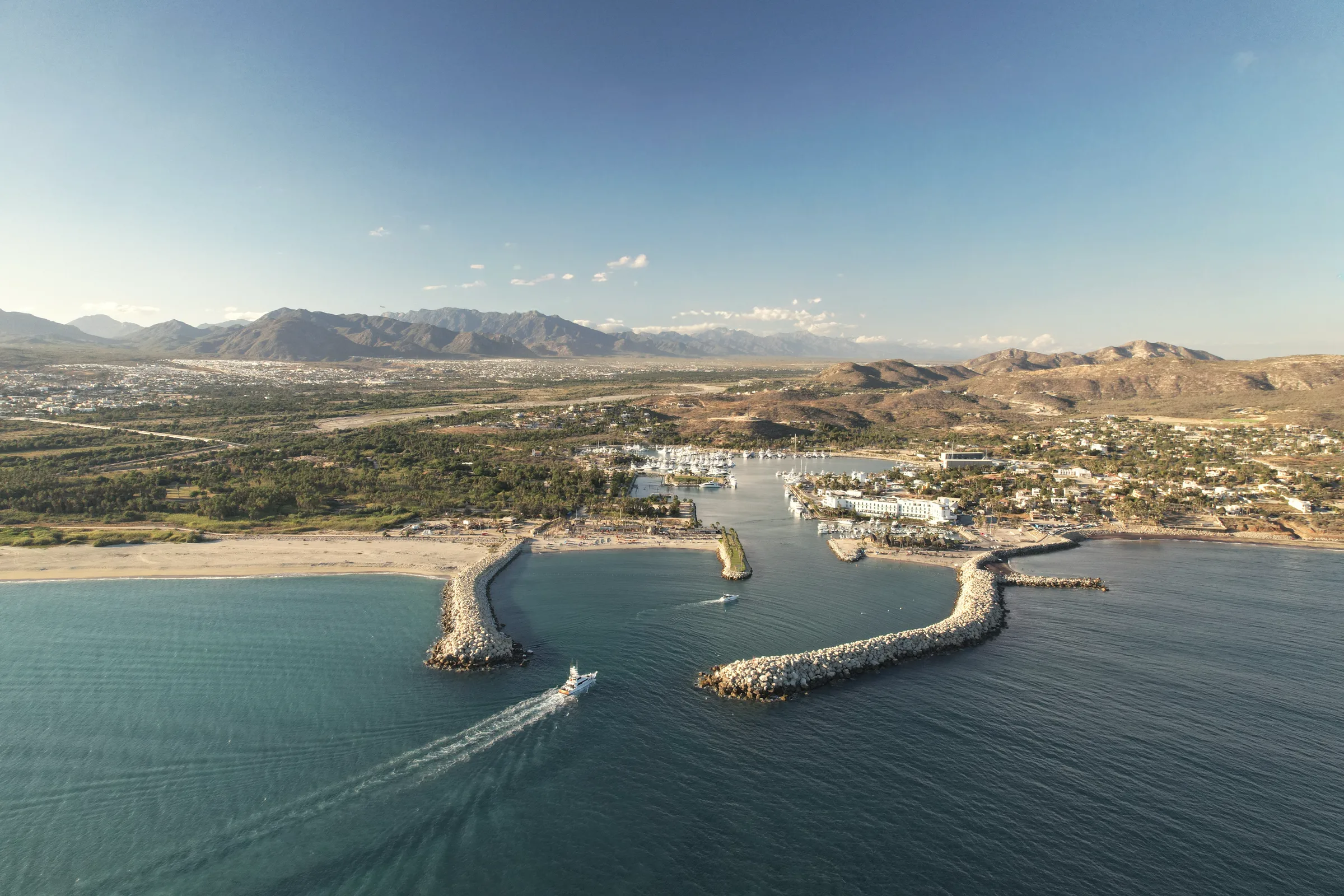 Aerial view of the Puerto Los Cabos marina entrance between two rock breakwaters, with the beach, the town and mountains behind.