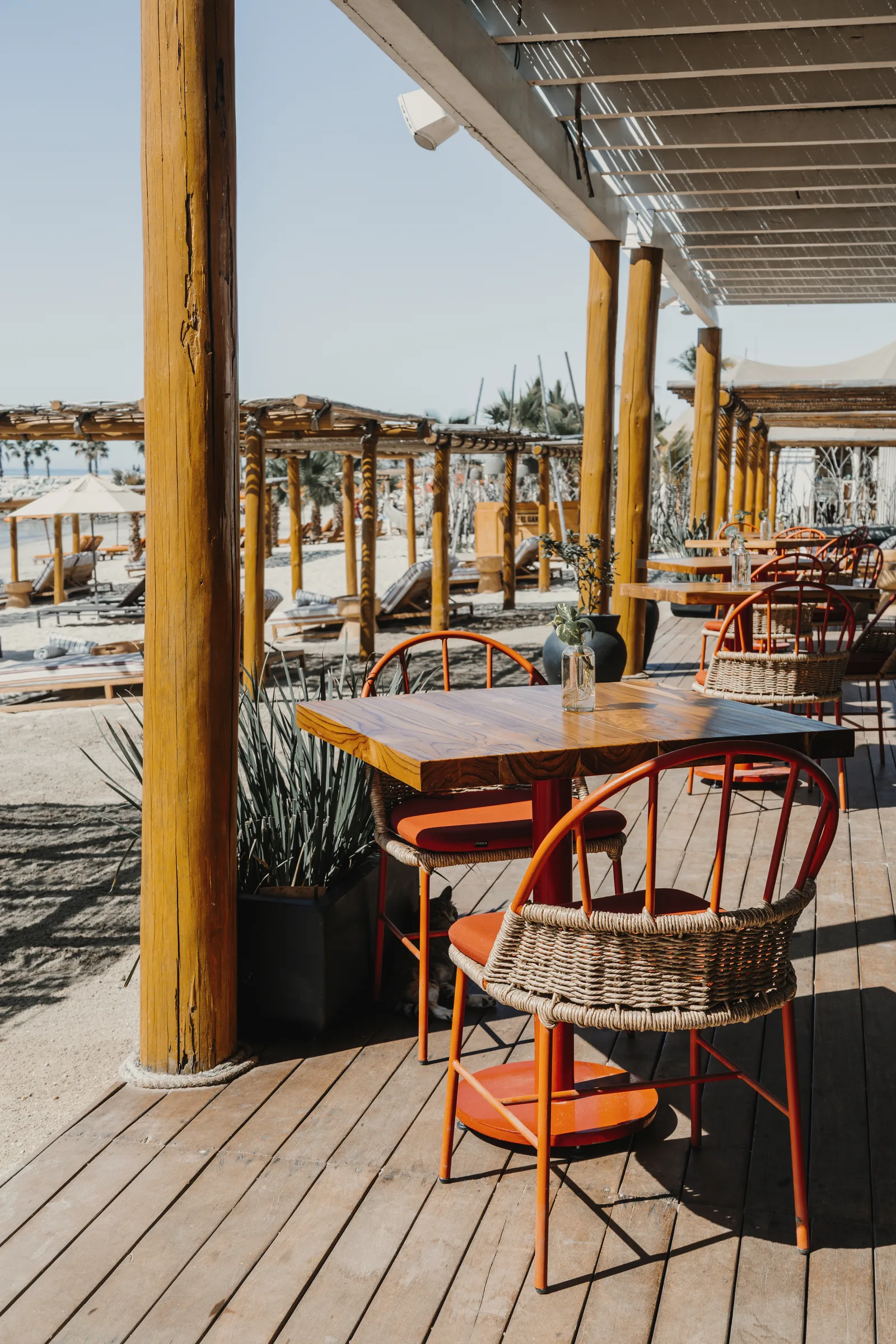 Covered wooden deck with wooden tables and orange chairs, looking out over beach loungers under shade structures toward the sea.