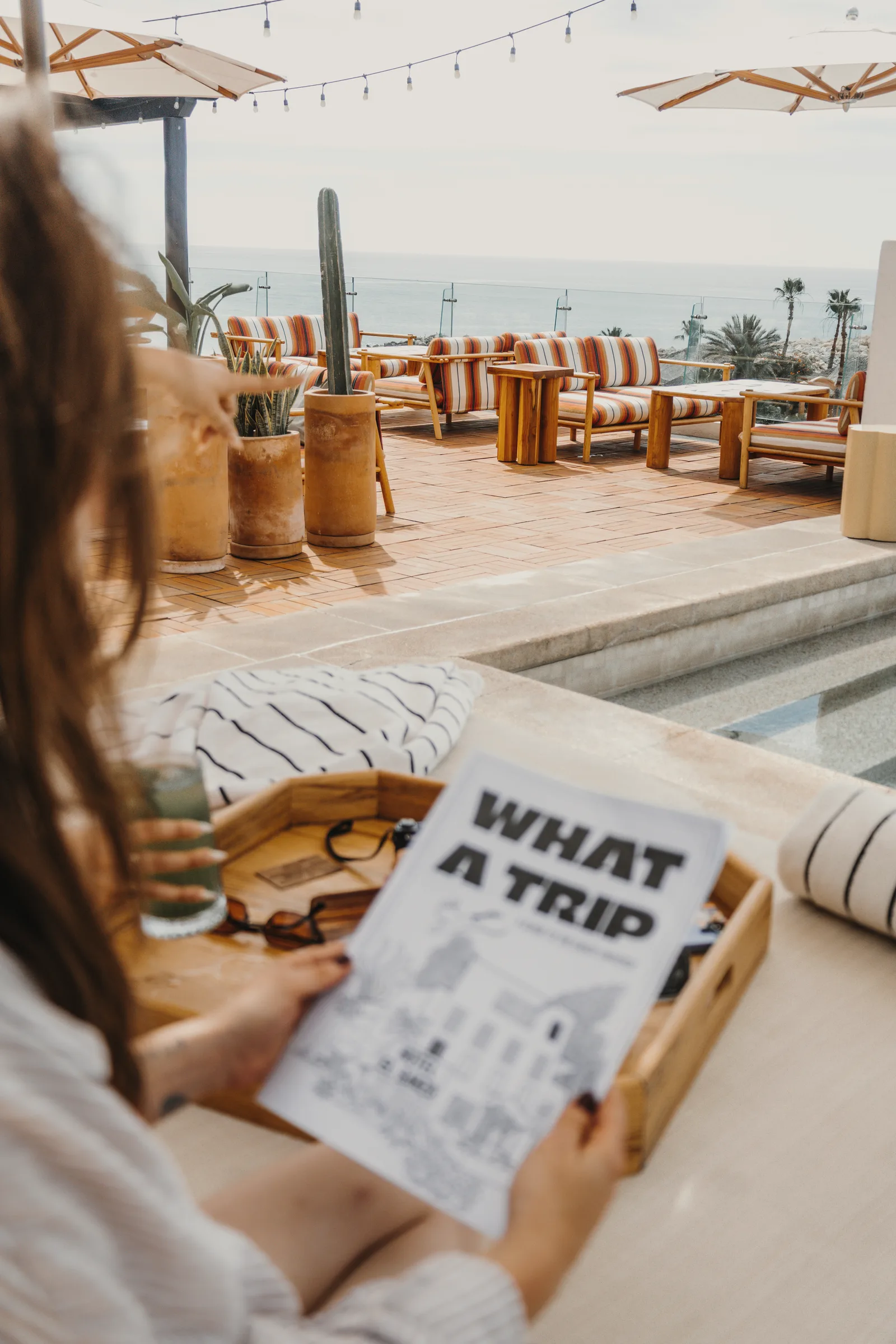 Guest holding a printed 'What a Trip' guide on a terrace with striped lounge seating, umbrellas and the sea in the background.