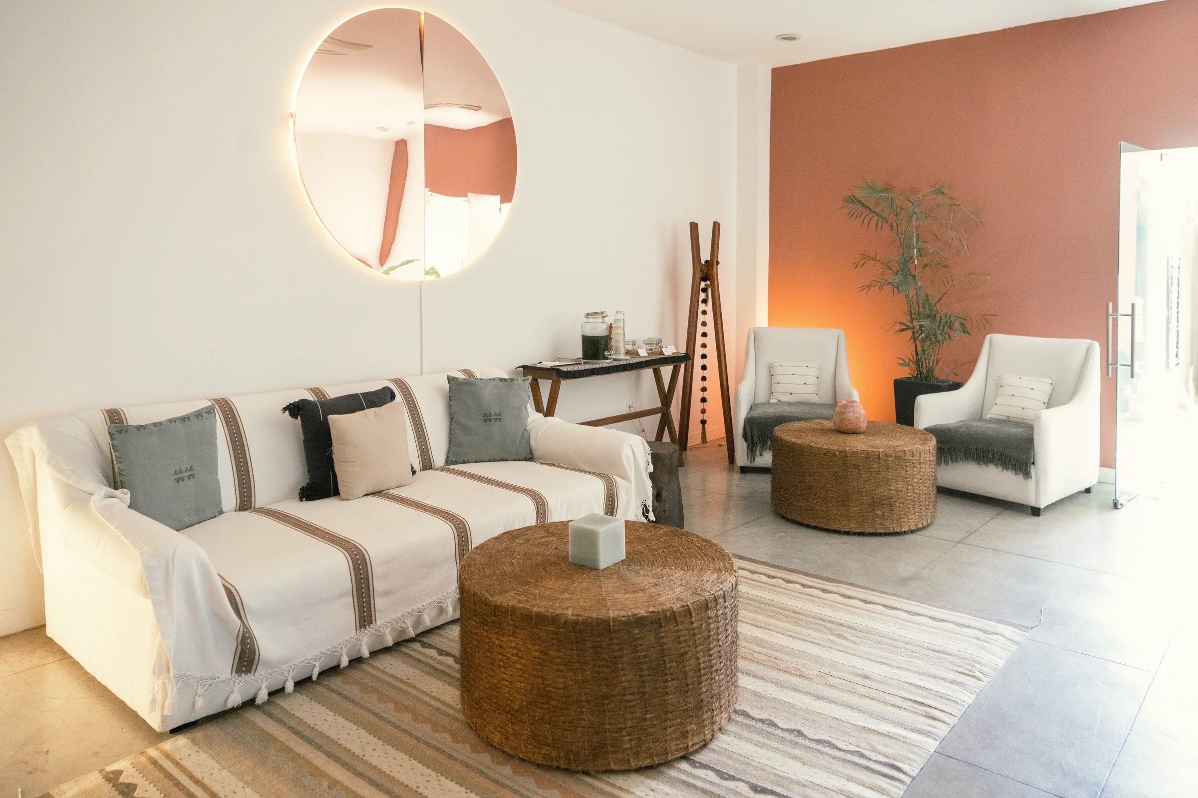 Lounge area with a white sofa, woven round tables and armchairs against a terracotta wall, with a round mirror.