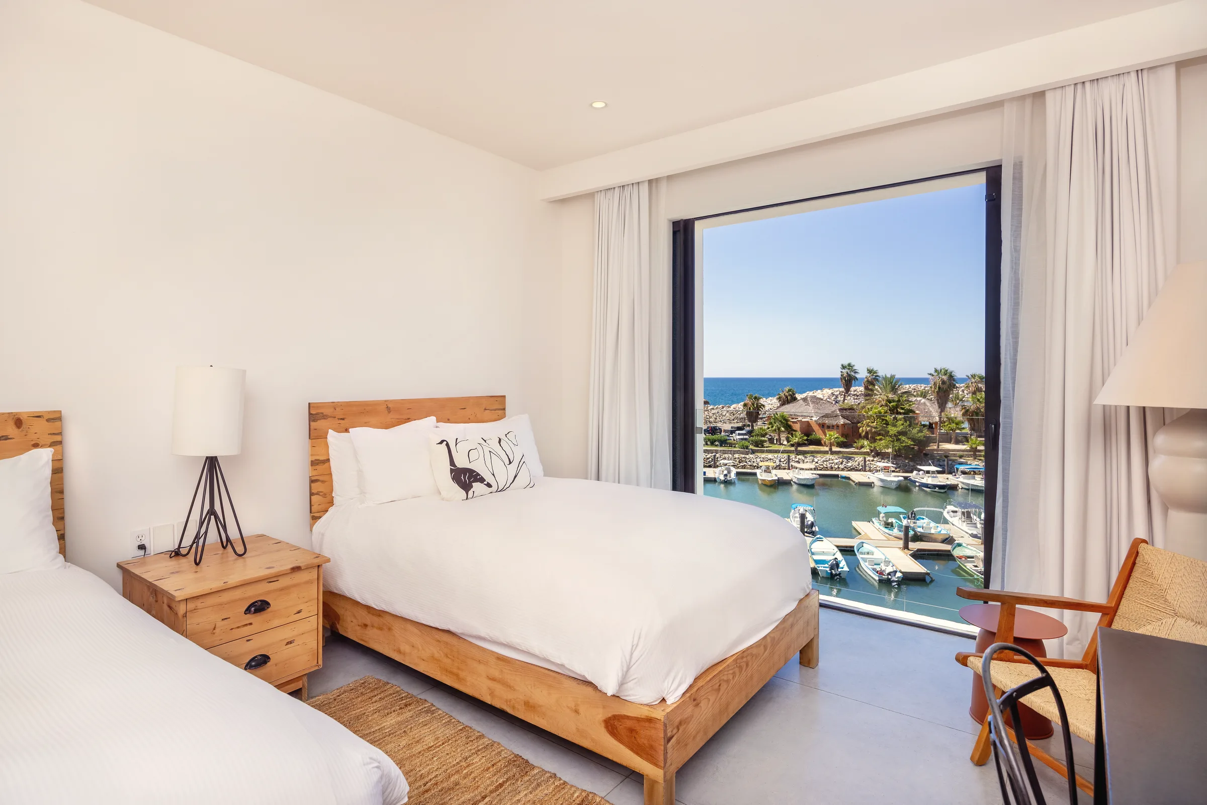 Two beds with wooden headboards and white linens beside a floor-to-ceiling window framed by sheer curtains, looking out over a marina with docked boats, palms and the sea in the distance.