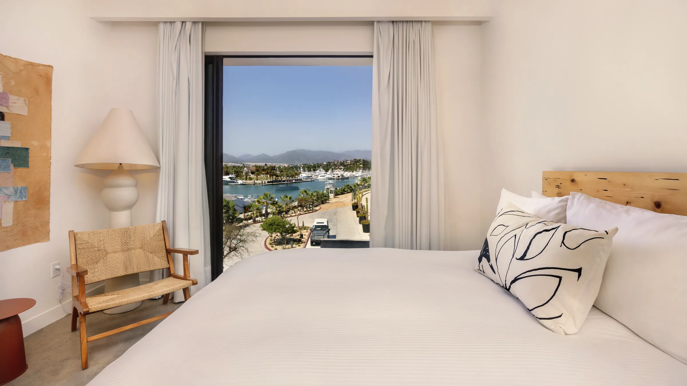 Bed with white linens beside a tall window with curtains, framing a view over the marina, palms and distant hills.
