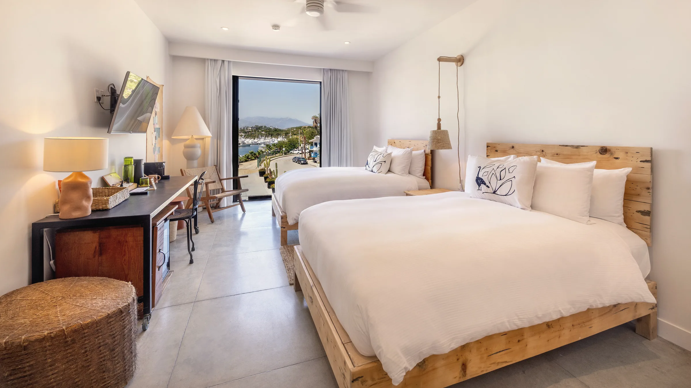Long view along the room past two beds with white linens, a desk and a television, toward a large window overlooking the marina.