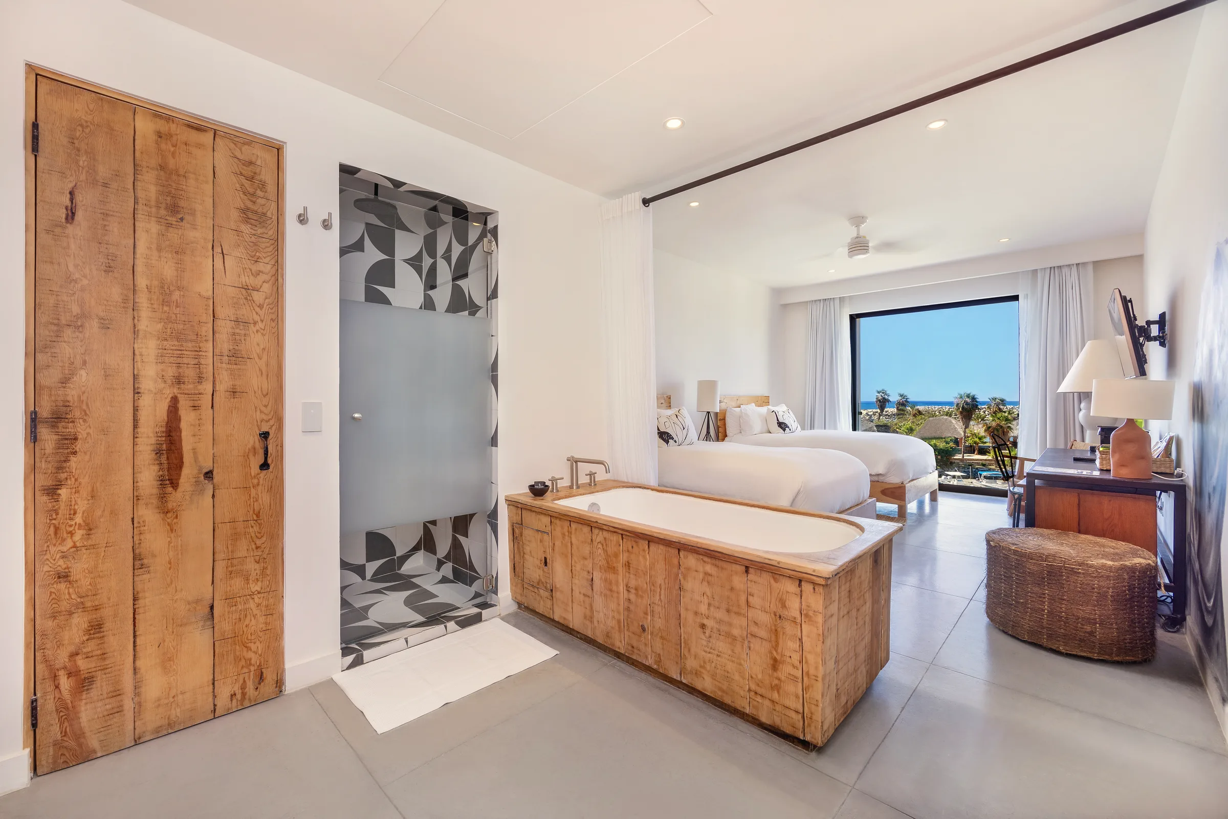 Bathroom with a wooden soaking tub, a tall wooden door and a patterned-tile shower, with the two beds and a large window visible beyond.