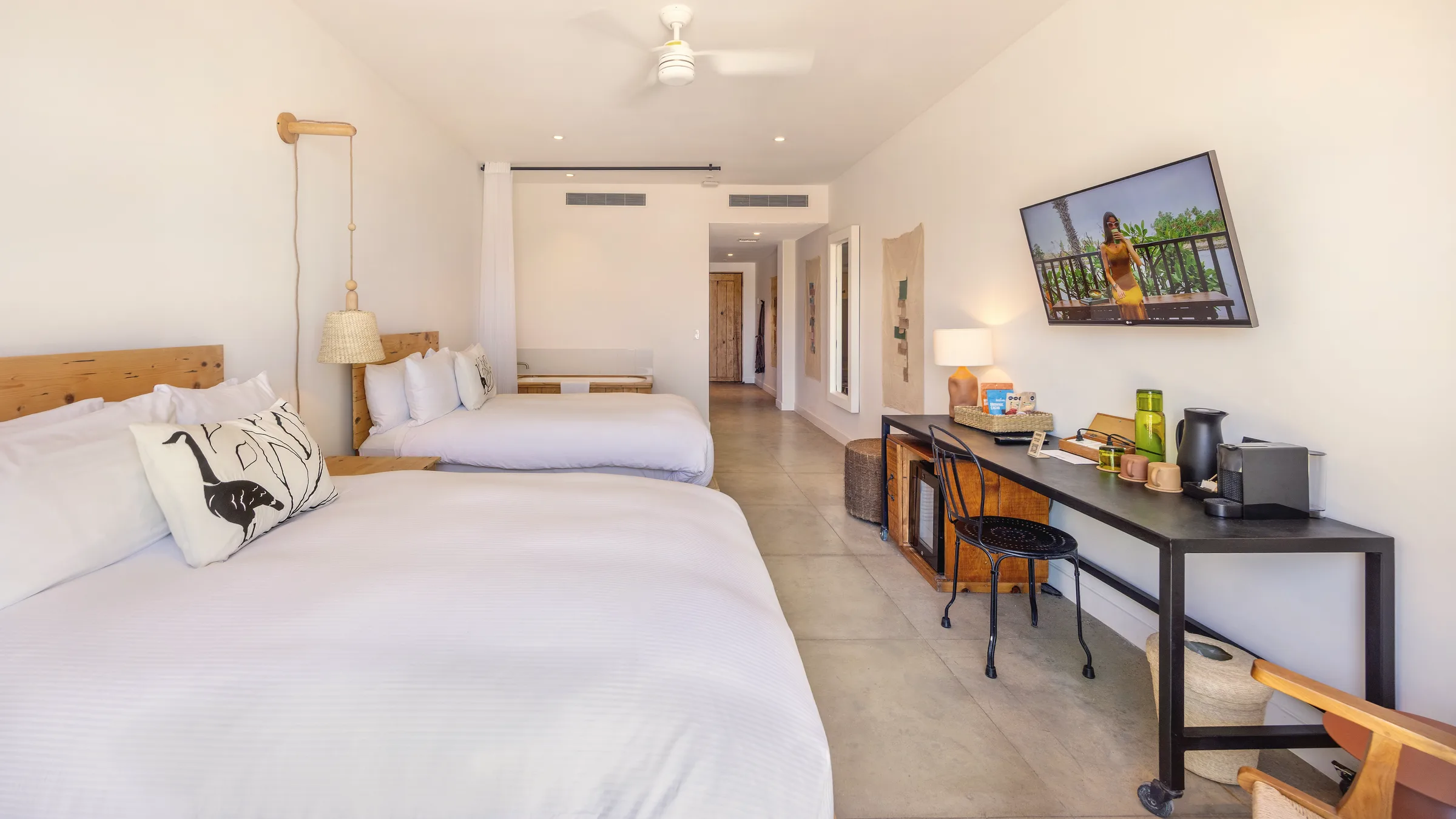 View down the length of the room from beside one bed, with a second bed, a black desk and a television, and the entry corridor at the far end.