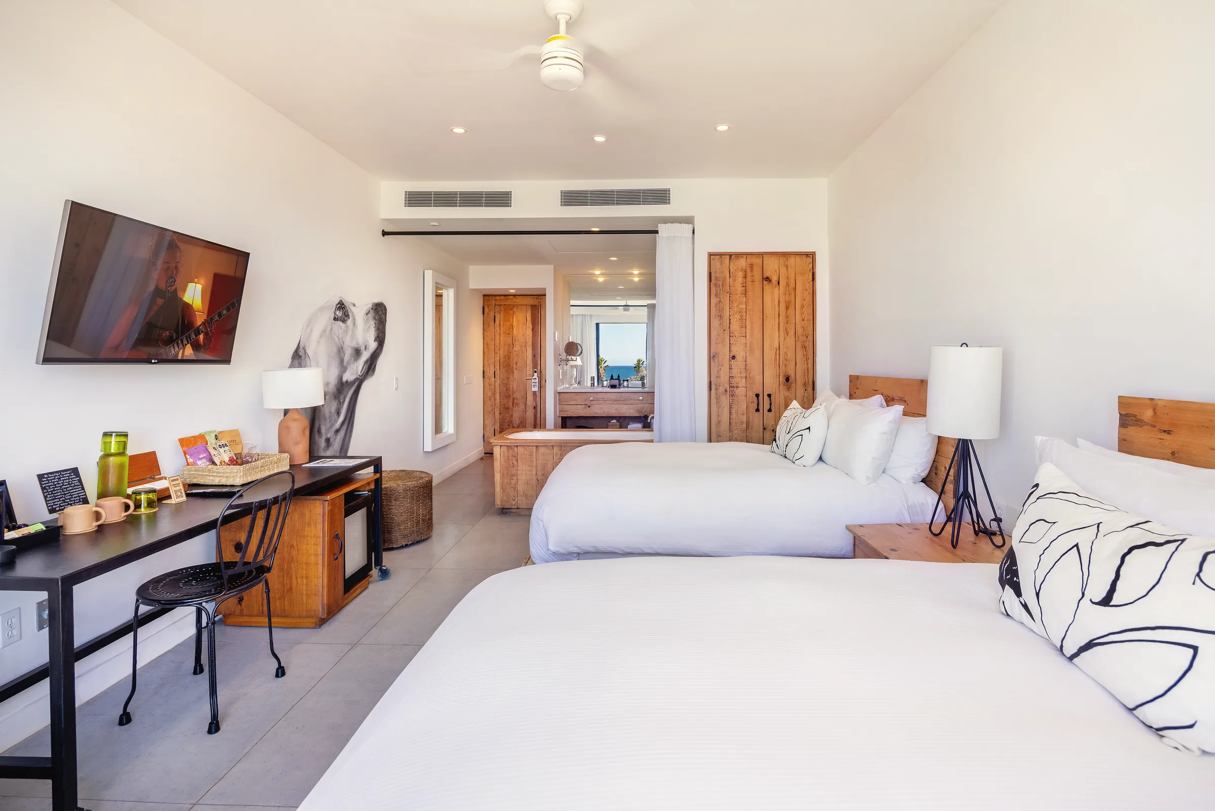 Two beds with white linens, a desk with a television and a ceiling fan, and the bathroom area with a soaking tub visible at the back.