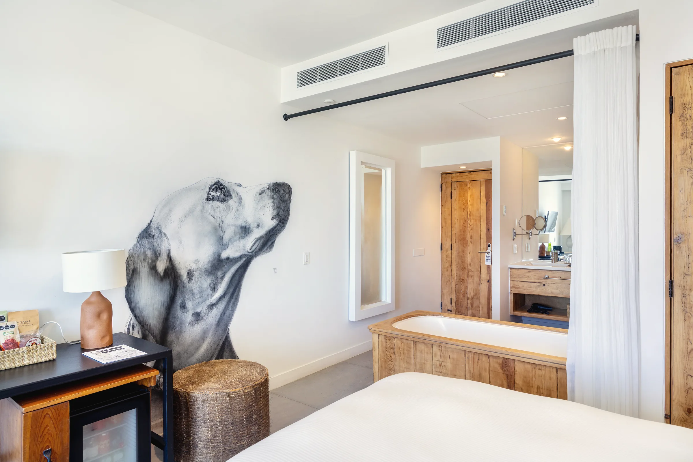 Bedroom wall with a large black-and-white dog mural, a table lamp and a woven stool, with a soaking tub and a curtain at the bathroom opening.