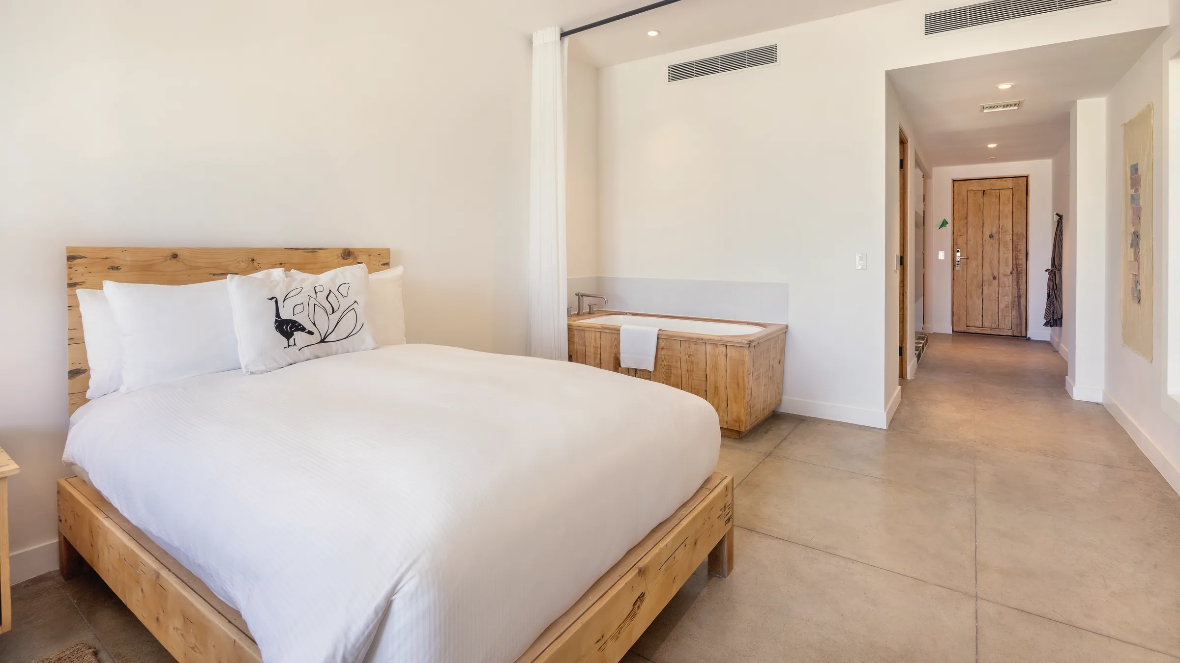 One of the beds, with white linens and a wooden headboard, in the foreground, with a wooden soaking tub and the entry corridor behind.