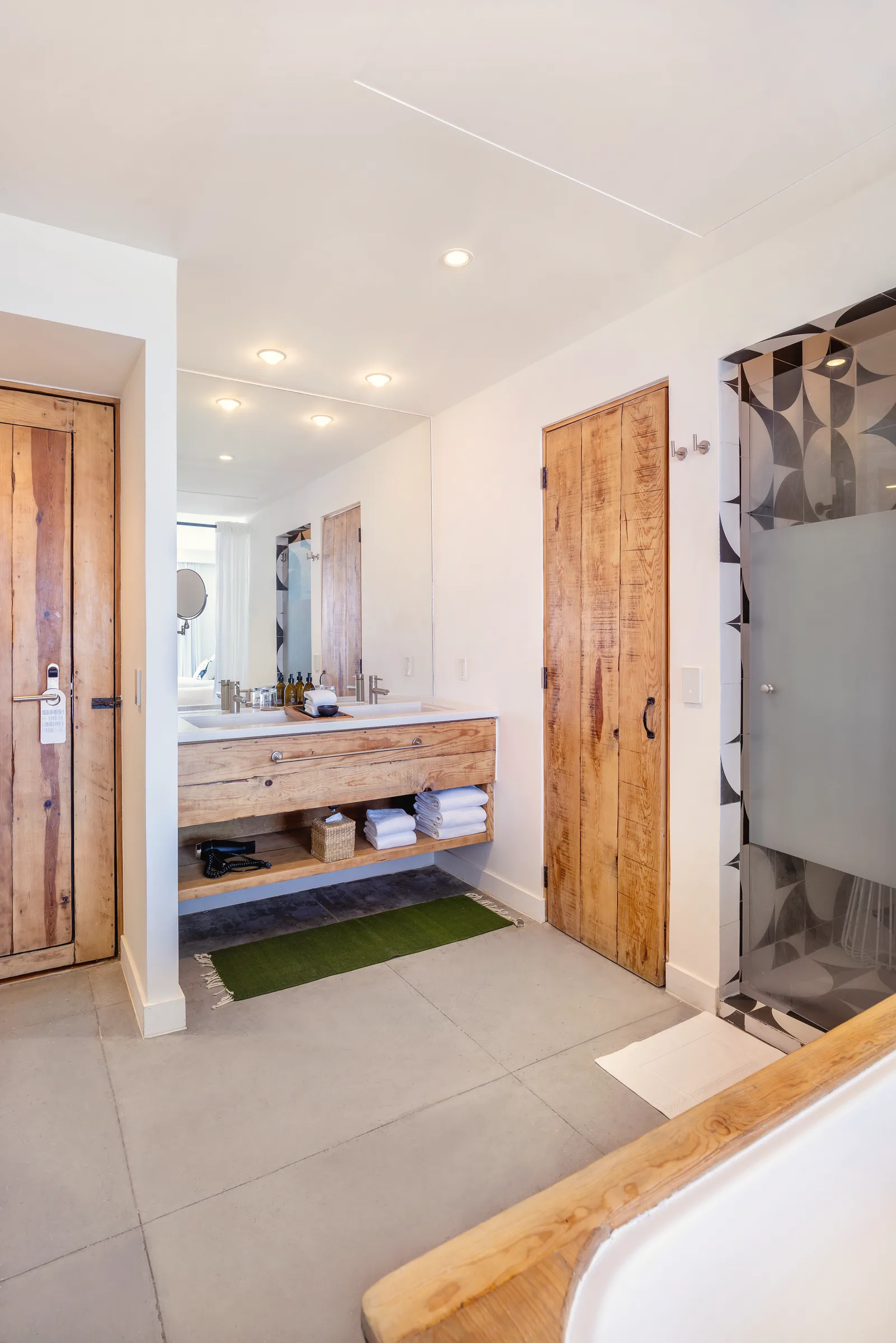 Wooden bathroom vanity with a white counter and an open shelf of folded towels, a mirror, wooden doors and the frosted-glass shower door.
