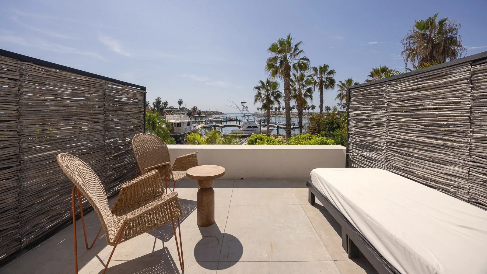 Private terrace enclosed by woven branch screens, with two wicker chairs, a small side table and a daybed, and palm trees and boats visible beyond the low wall.