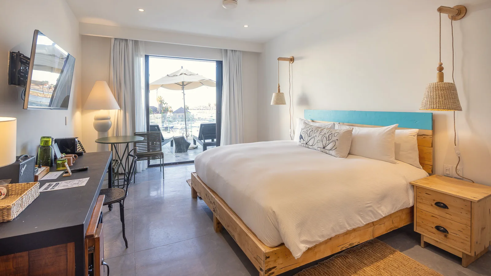 King bed with a turquoise headboard and a desk, with a sliding glass door opening to a terrace with a patio umbrella and the marina outside.