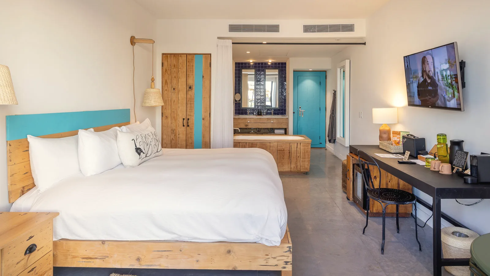 King bed with a turquoise headboard and white linens, with a desk and television on the right, and a wooden soaking tub, wardrobe and turquoise door behind.