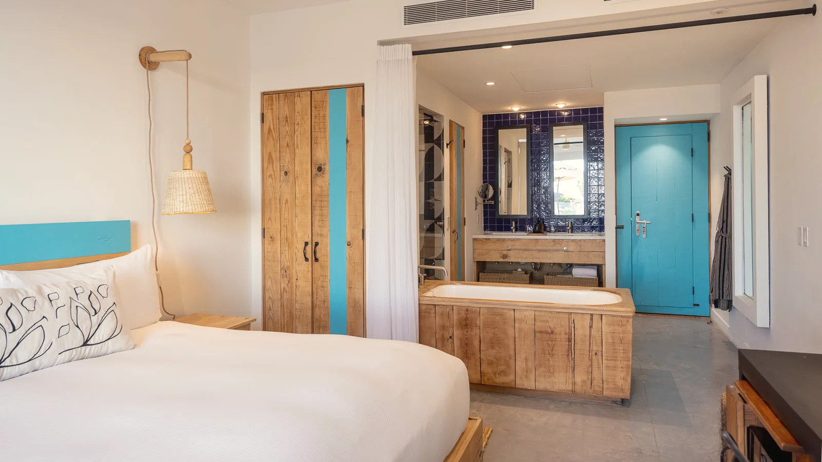 Corner of the king bed with a turquoise headboard in the foreground, and the soaking tub, vanity and turquoise entry door in the background.