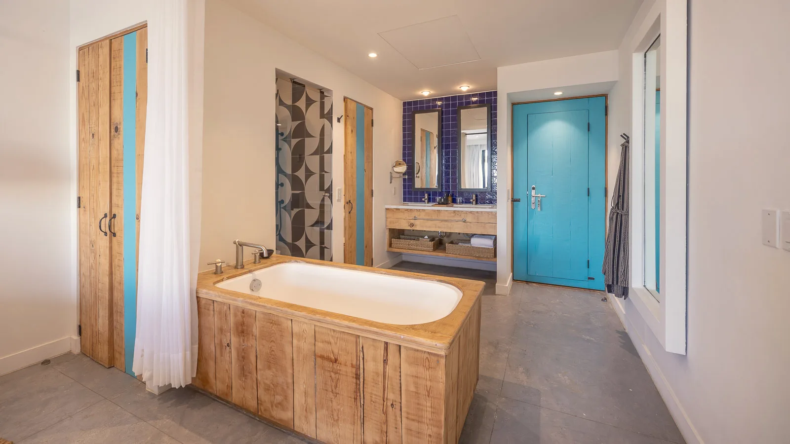 Bathroom area with a wooden soaking tub, a vanity with a blue-tiled backsplash, a wooden wardrobe, and the turquoise entry door.