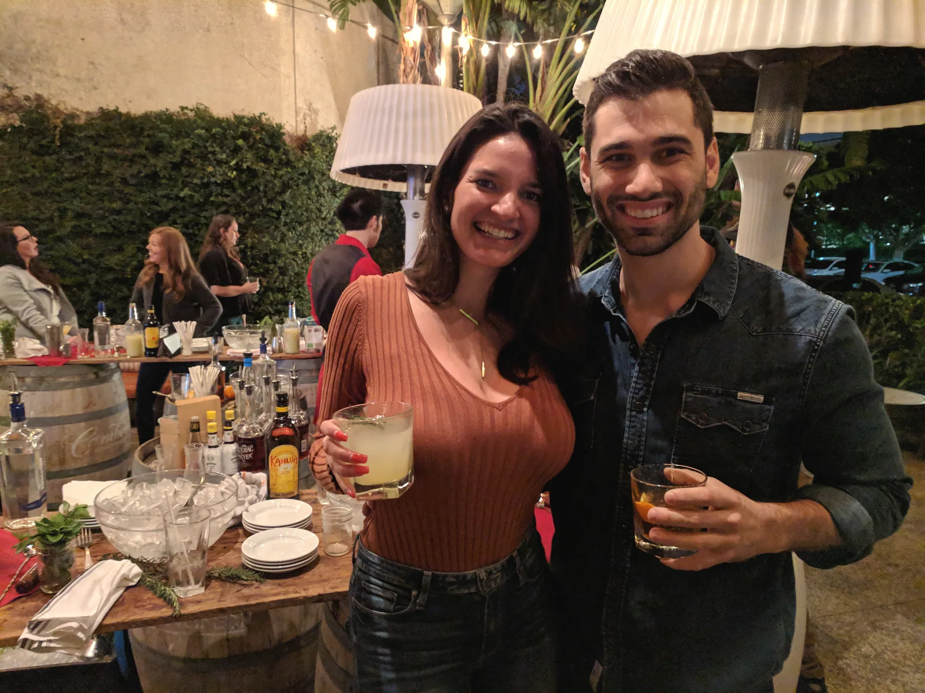 Carmela and Charlie each holding a drink at a party, a laden bar table and string lights behind them