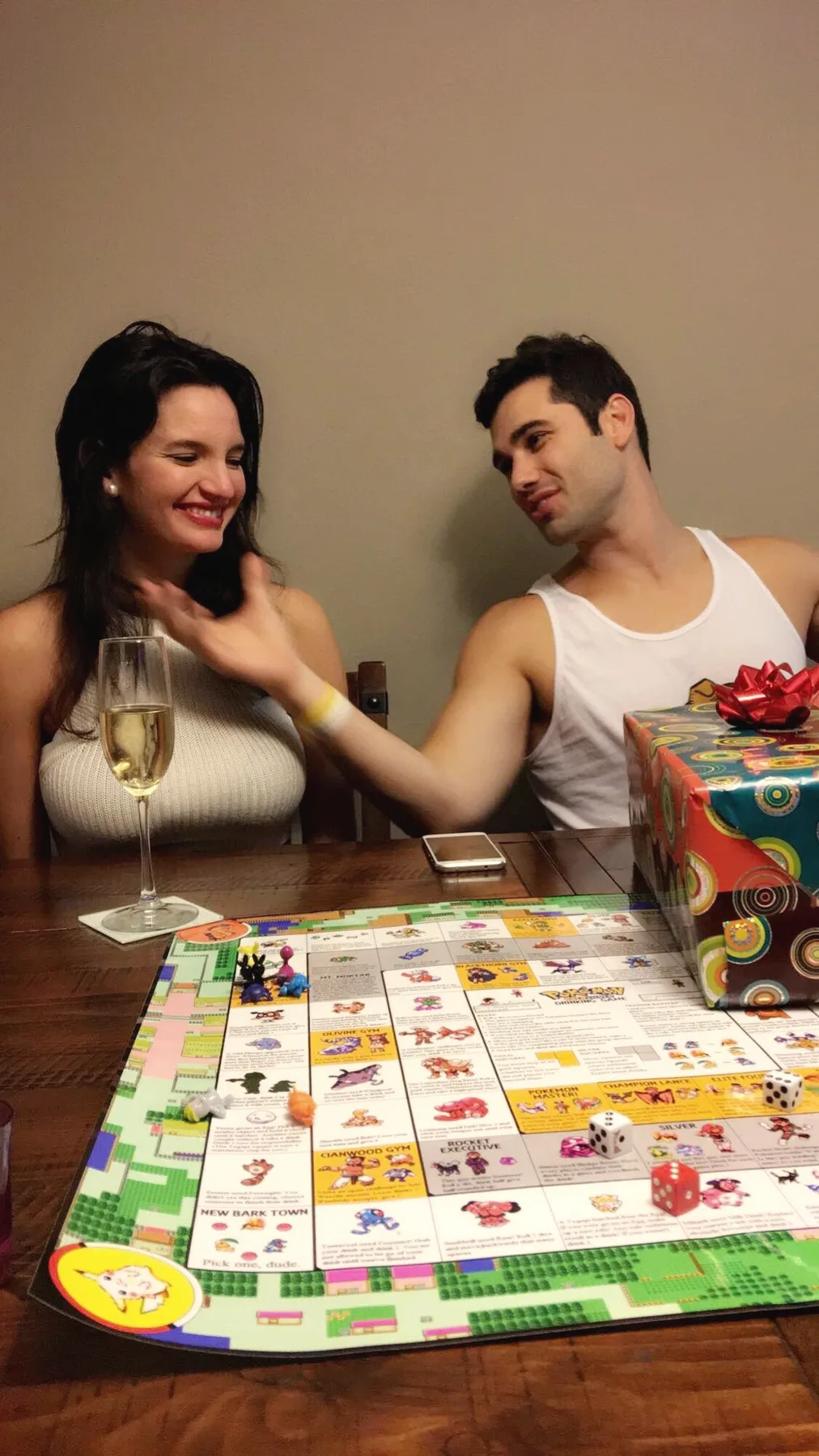 Carmela laughing with a glass of champagne while Charlie leans in beside a colourful board game on a wooden table
