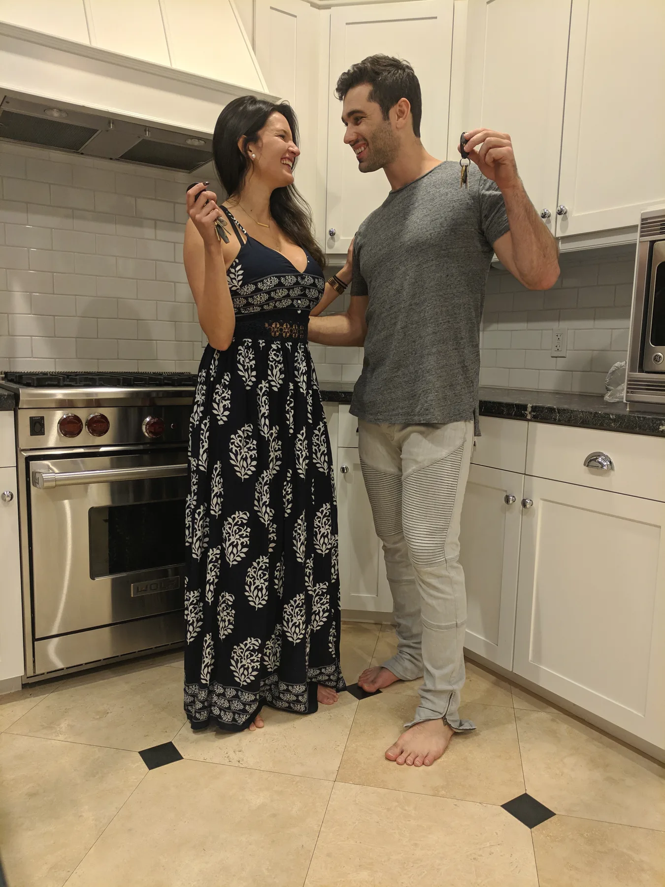 Carmela and Charlie in a bright white kitchen, each holding up a set of new keys and smiling at each other