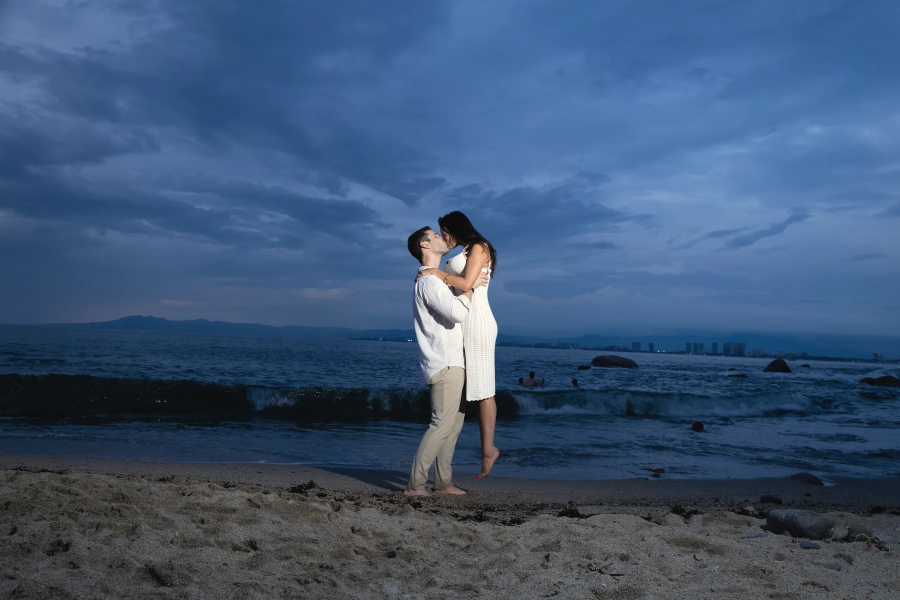 Carmela and Charlie embracing small on the sand under a deep blue evening sky, the ocean behind them
