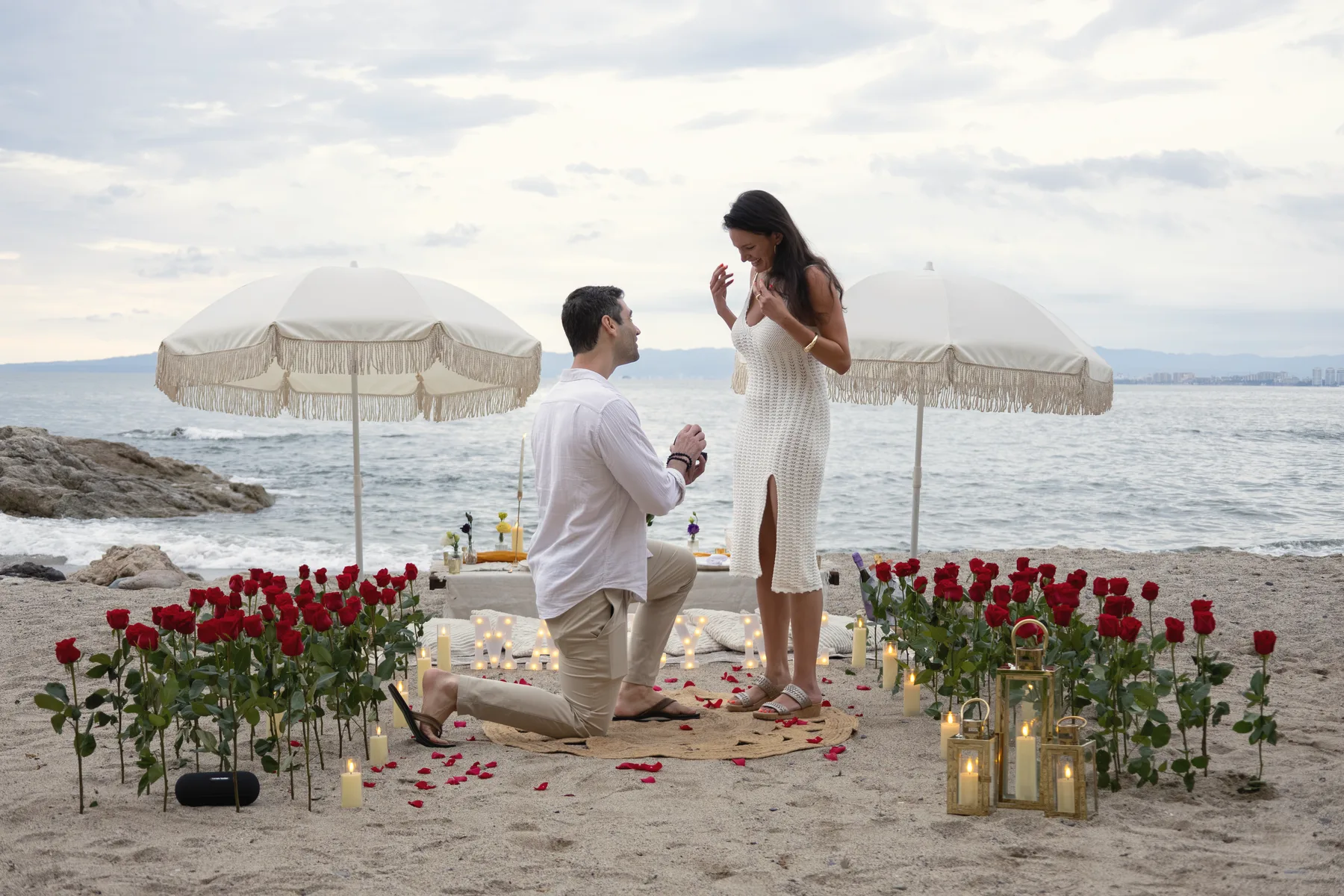 Charlie kneeling on the beach as Carmela covers her mouth, a ring of red roses, candles and two white fringed umbrellas around them by the sea