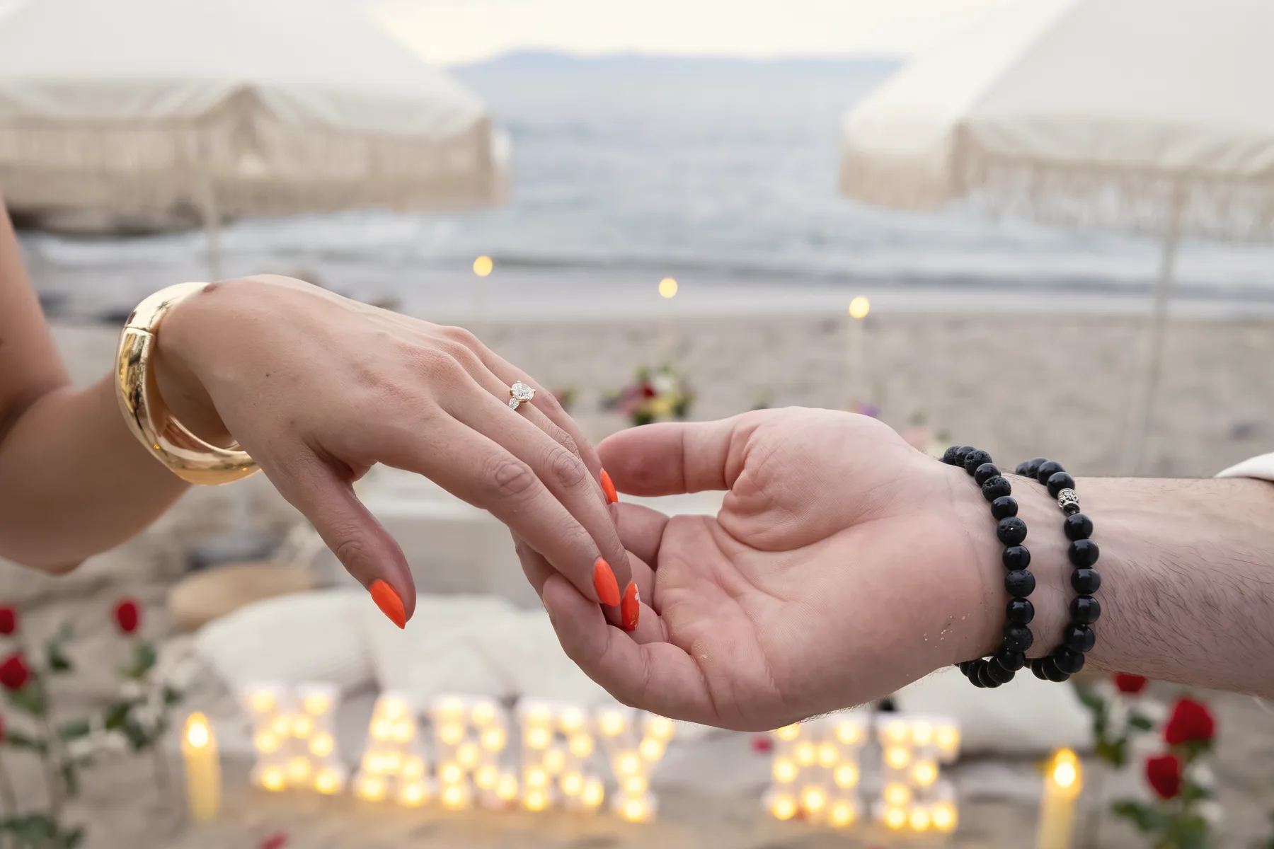 Carmela's hand with her engagement ring resting in Charlie's hand, a lit MARRY ME sign and rose petals on the sand behind