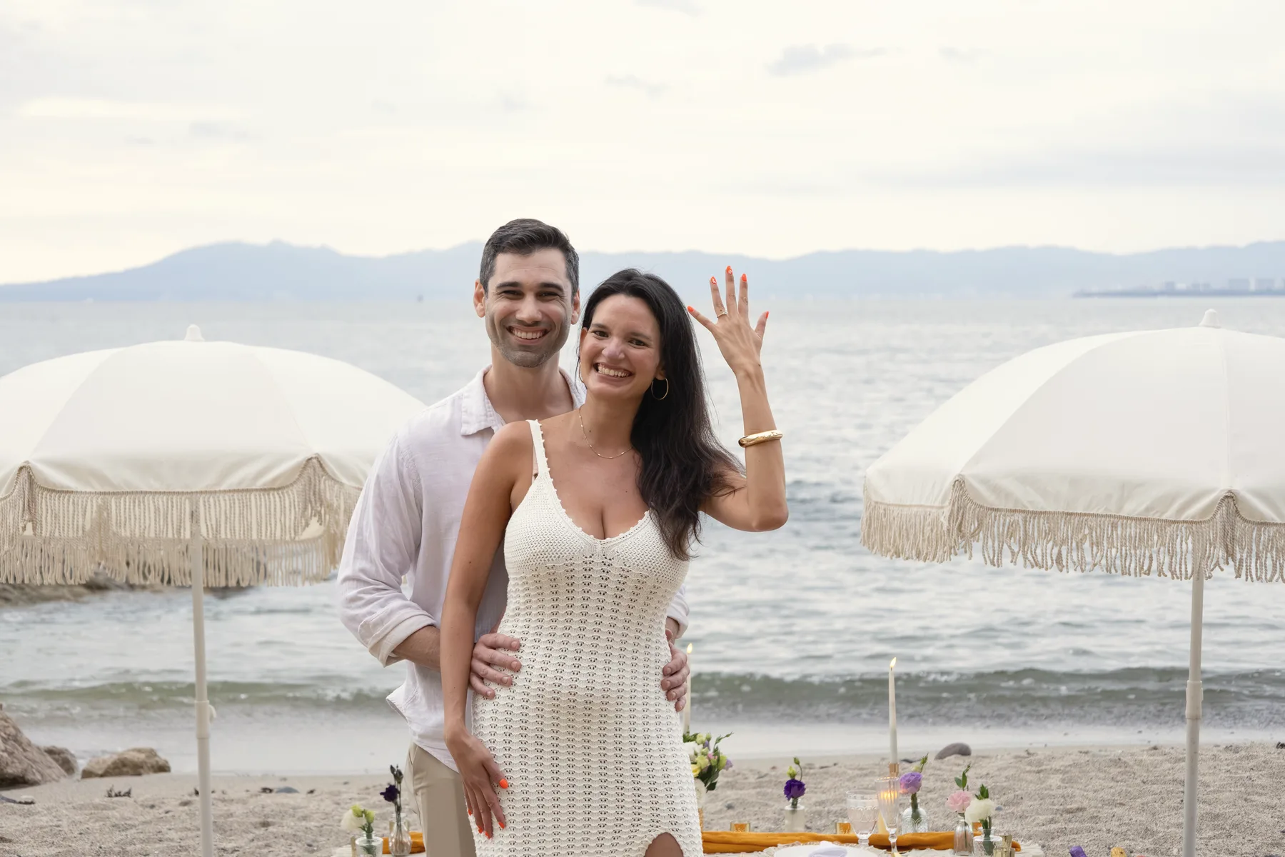 Charlie standing behind Carmela on the beach beside white fringed umbrellas as she lifts a hand, the sea and hazy mountains beyond