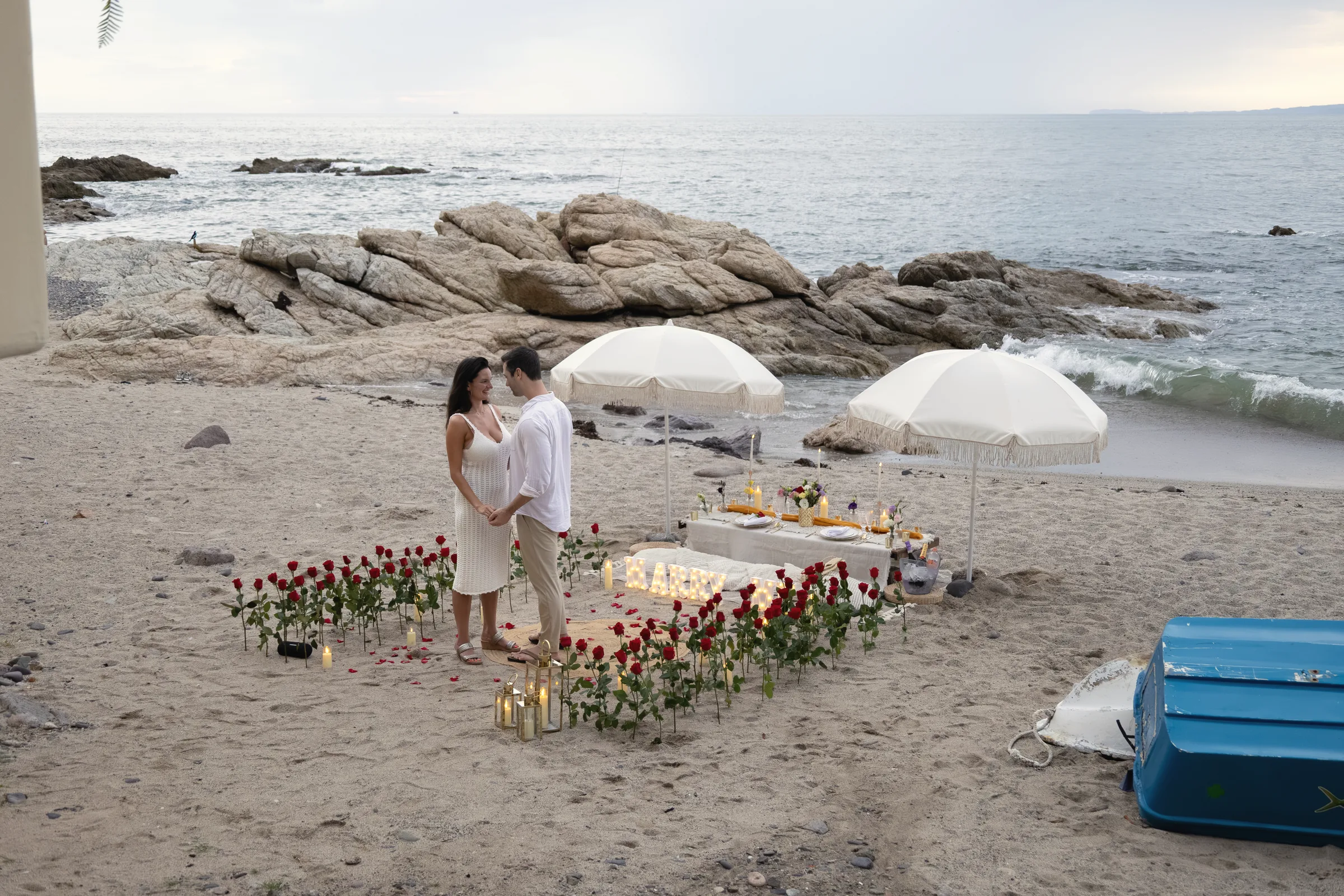 A candlelit table ringed by red roses on a rocky beach, white fringed umbrellas and the sea behind, Carmela and Charlie standing beside it
