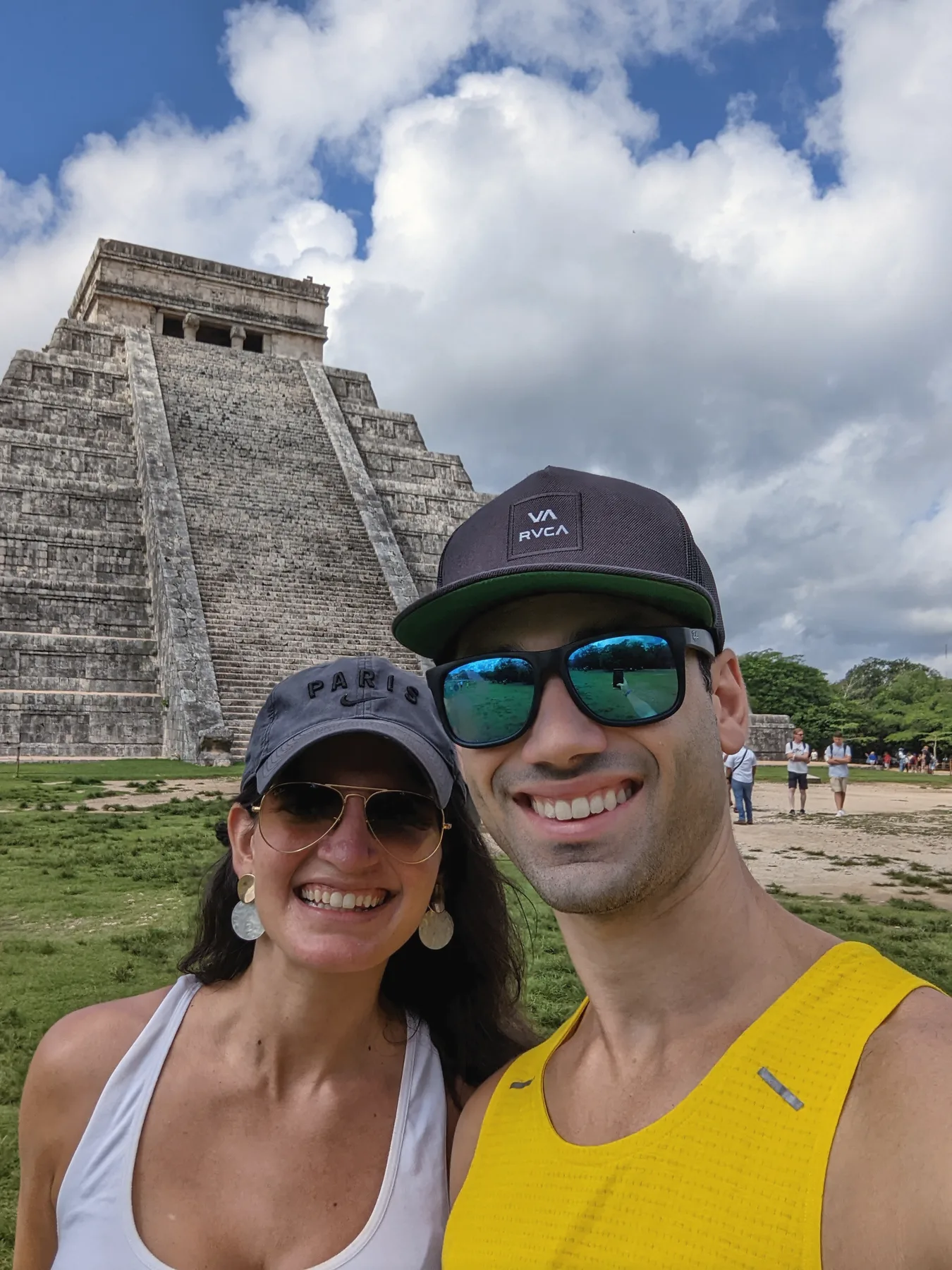 Carmela and Charlie in caps and sunglasses, smiling, with a tall stepped stone pyramid and big clouds behind them