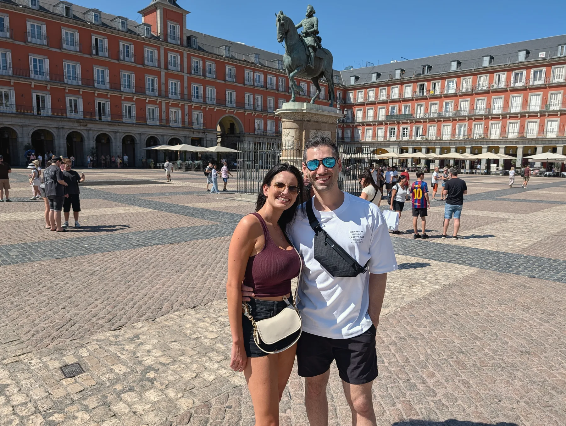Carmela and Charlie posing in a large cobbled square with red-brick arcaded buildings and a statue on a horse behind them