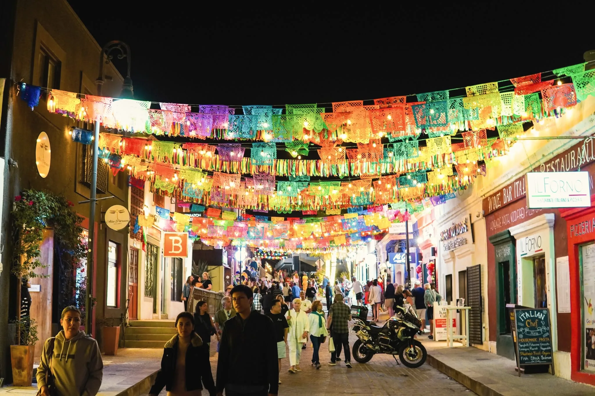 Busy street at night under strings of colourful papel picado banners, with shopfronts on both sides