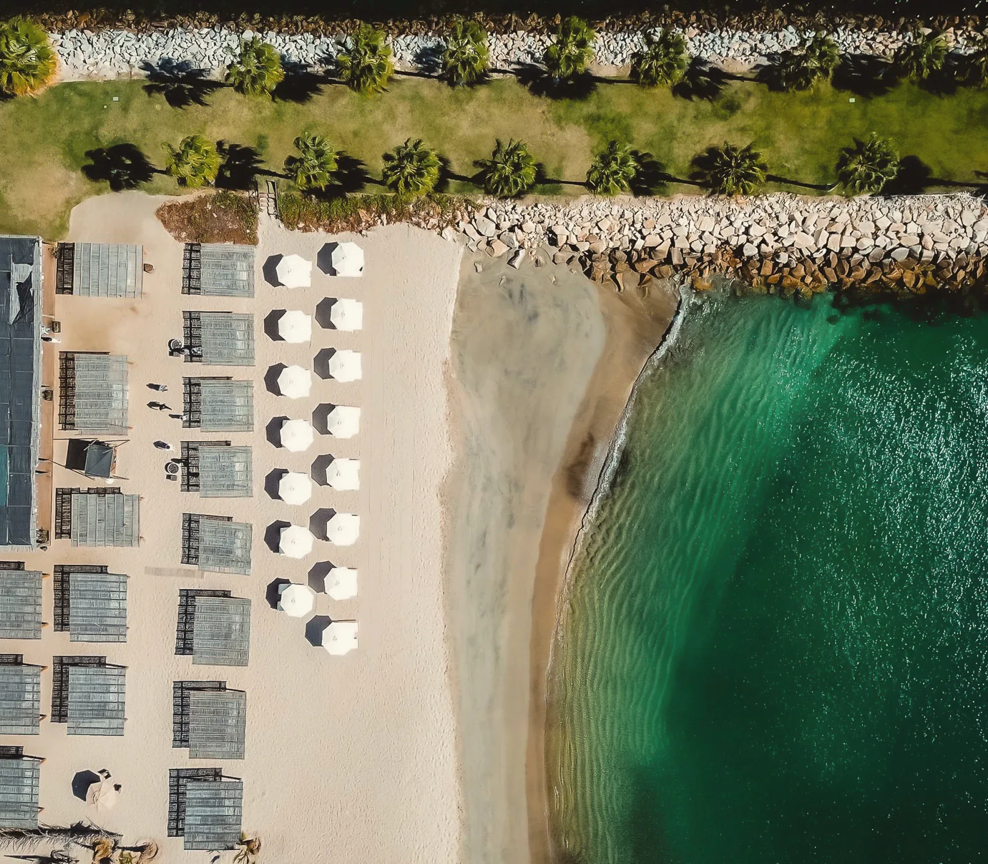 Aerial view of a beach club: rows of white umbrellas and cabanas on the sand beside green-blue water and a rock breakwater