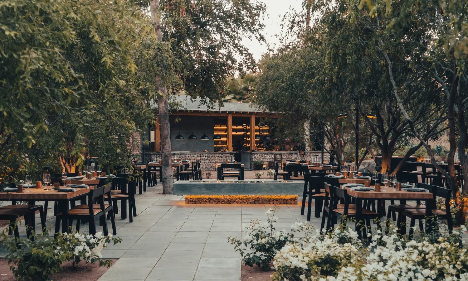 Open-air restaurant courtyard among trees, with rows of dark wooden tables on a paved floor and a low building behind