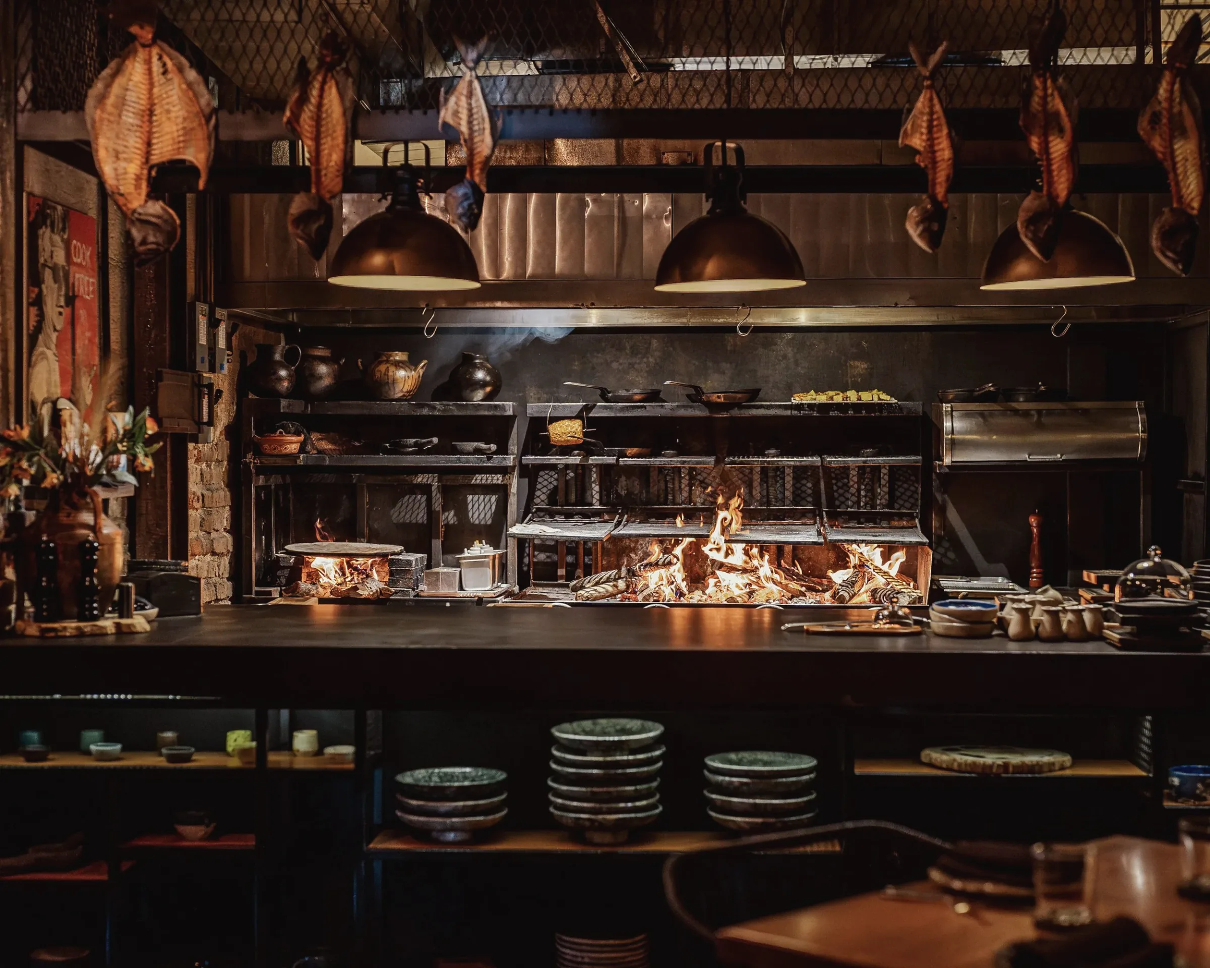 Open-fire kitchen with a wood fire burning under a grill, pendant lamps overhead and stacked plates below