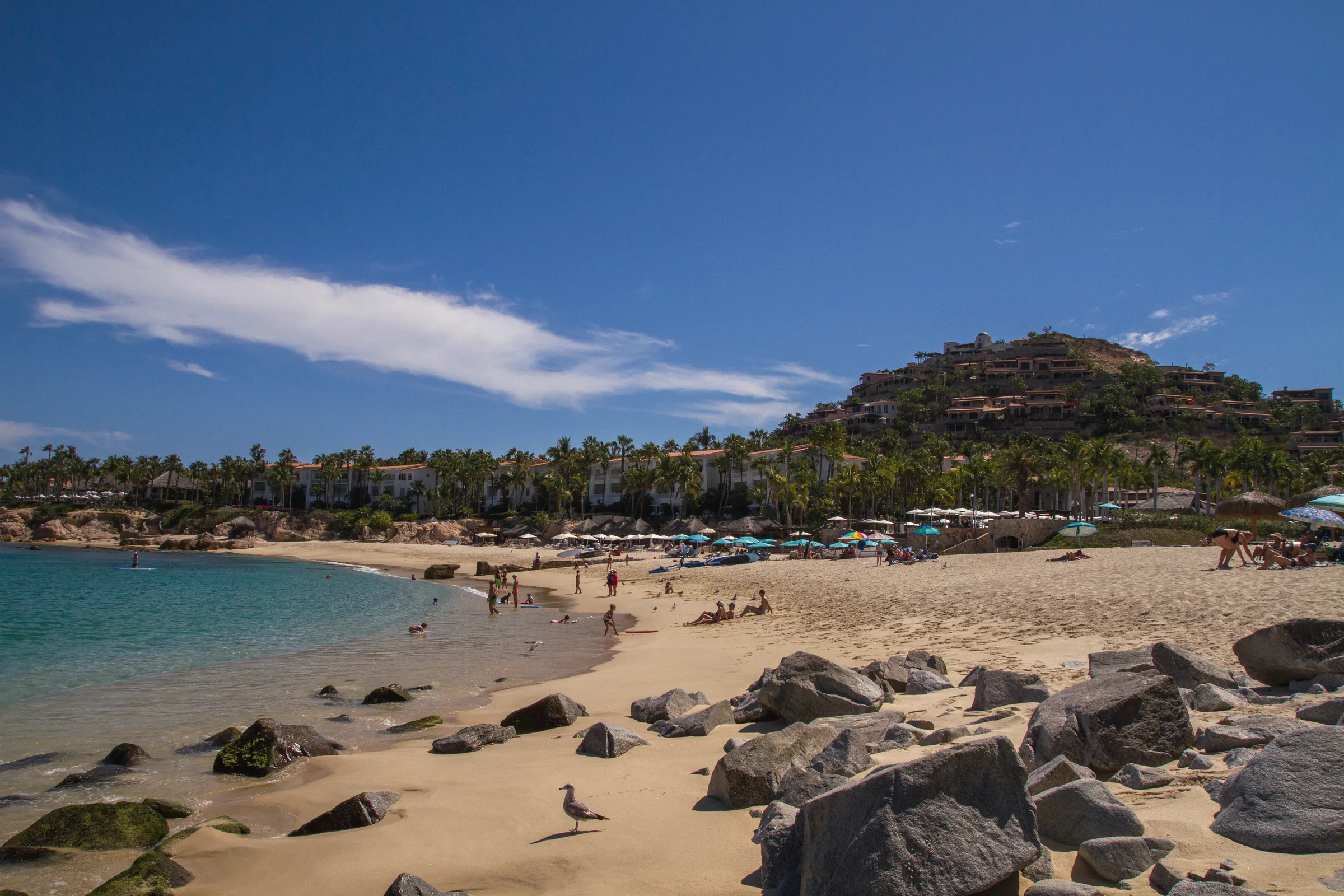 Sandy cove with rocks in the foreground, calm turquoise water and a hillside of resort buildings under a deep blue sky