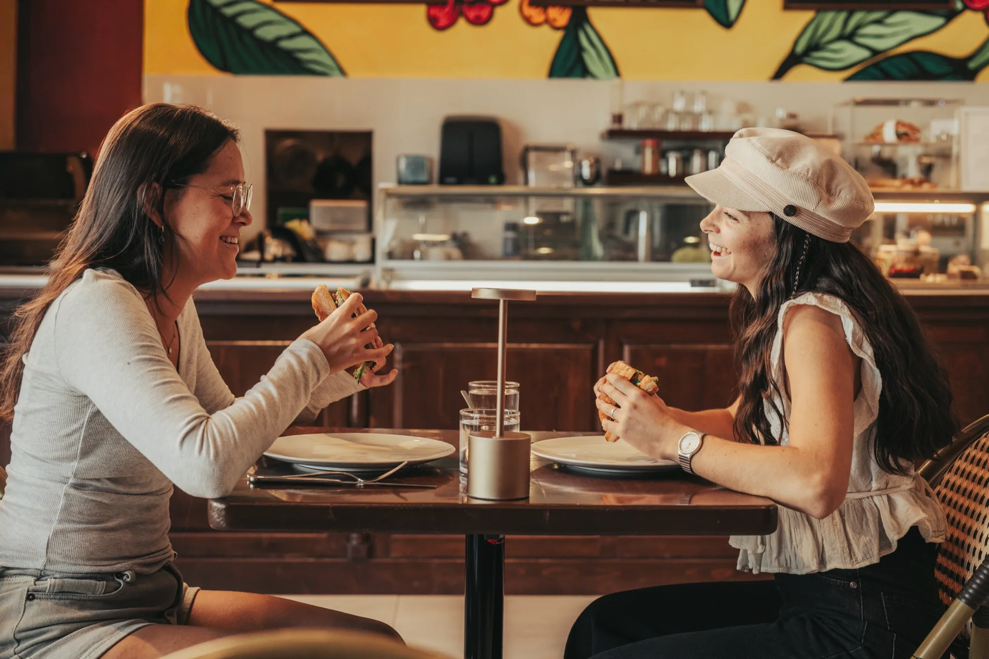 Two women laughing over sandwiches at a café table, a colourful mural and a pastry counter behind them