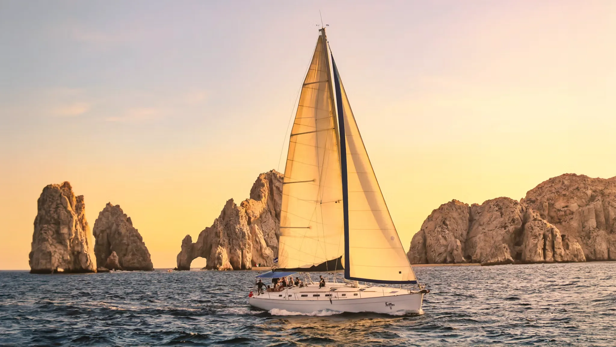 Sailboat under full sail passing rock formations at sunset, the sky glowing orange and pink